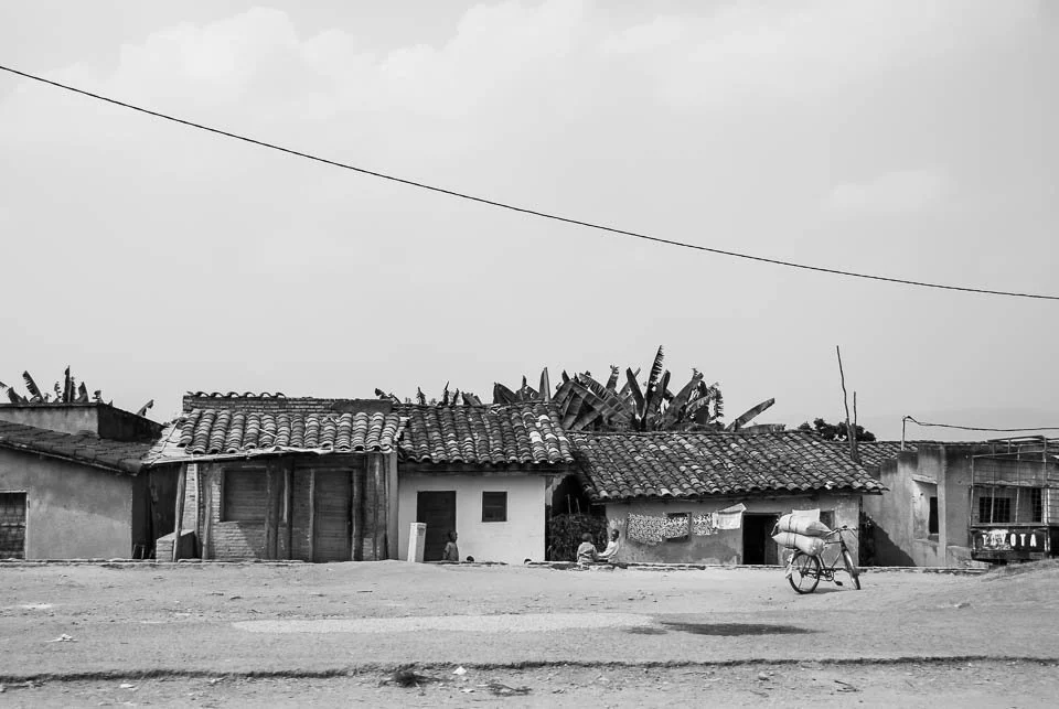 A black and white photo of small, rustic houses with tiled and corrugated metal roofs, along a dirt road, with a person pushing a bicycle loaded with bags in front.