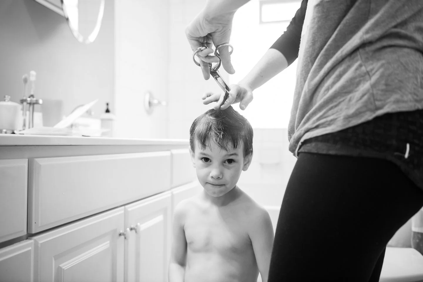 A young boy with wet hair standing in a bathroom as an adult cuts his hair with scissors. The bathroom has a sink and cabinets.