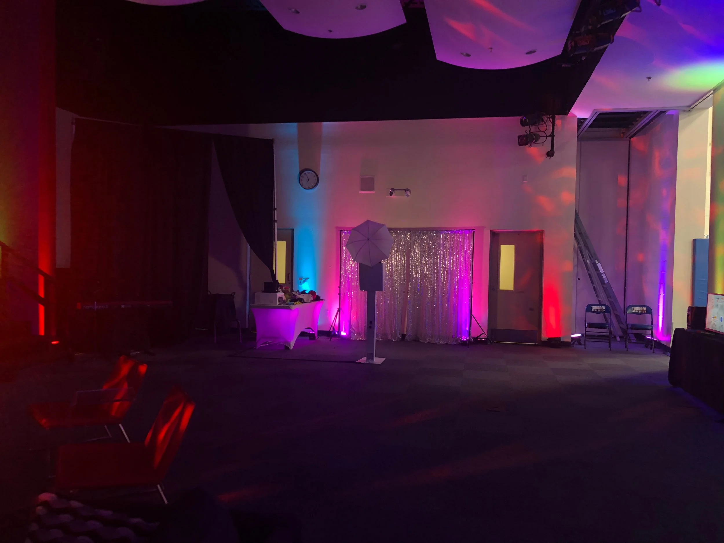Empty event space with colorful lighting, a photo booth with a sparkling backdrop, chairs, and a ladder, suggesting preparations for a celebration or party.