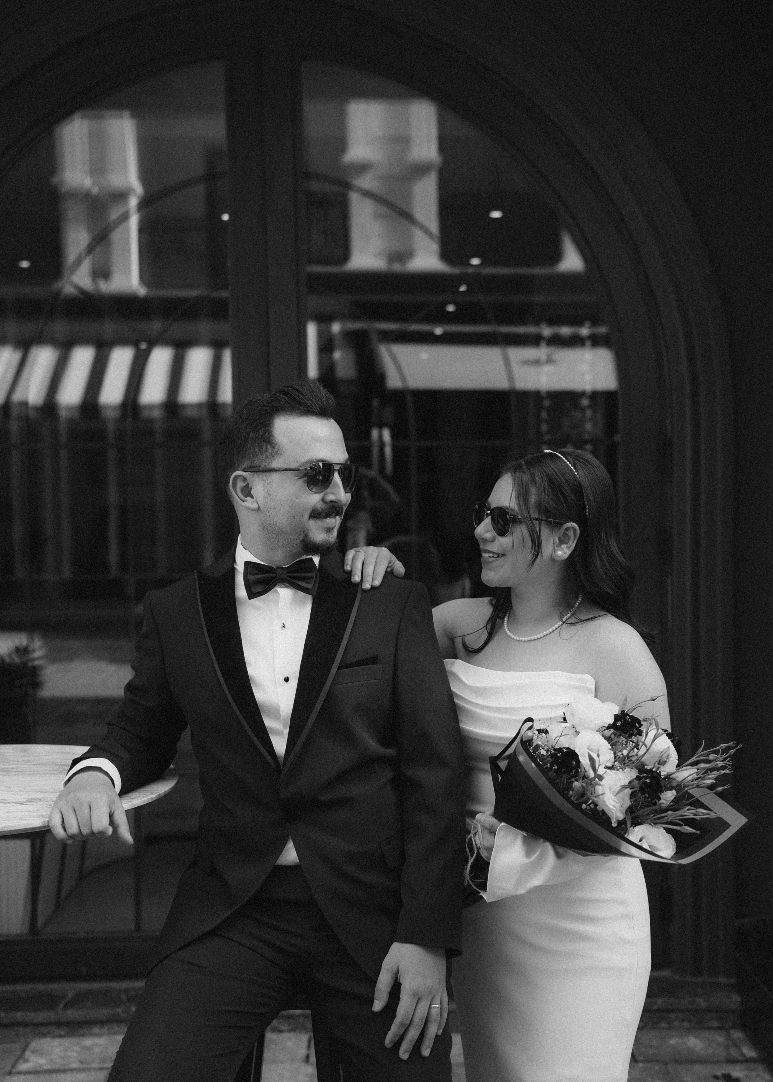 A black and white photo of a man and woman in formal attire, both wearing sunglasses. The man is in a tuxedo with a bow tie, leaning against a table, while the woman is in a strapless dress, holding a bouquet of flowers, and smiling at the man.