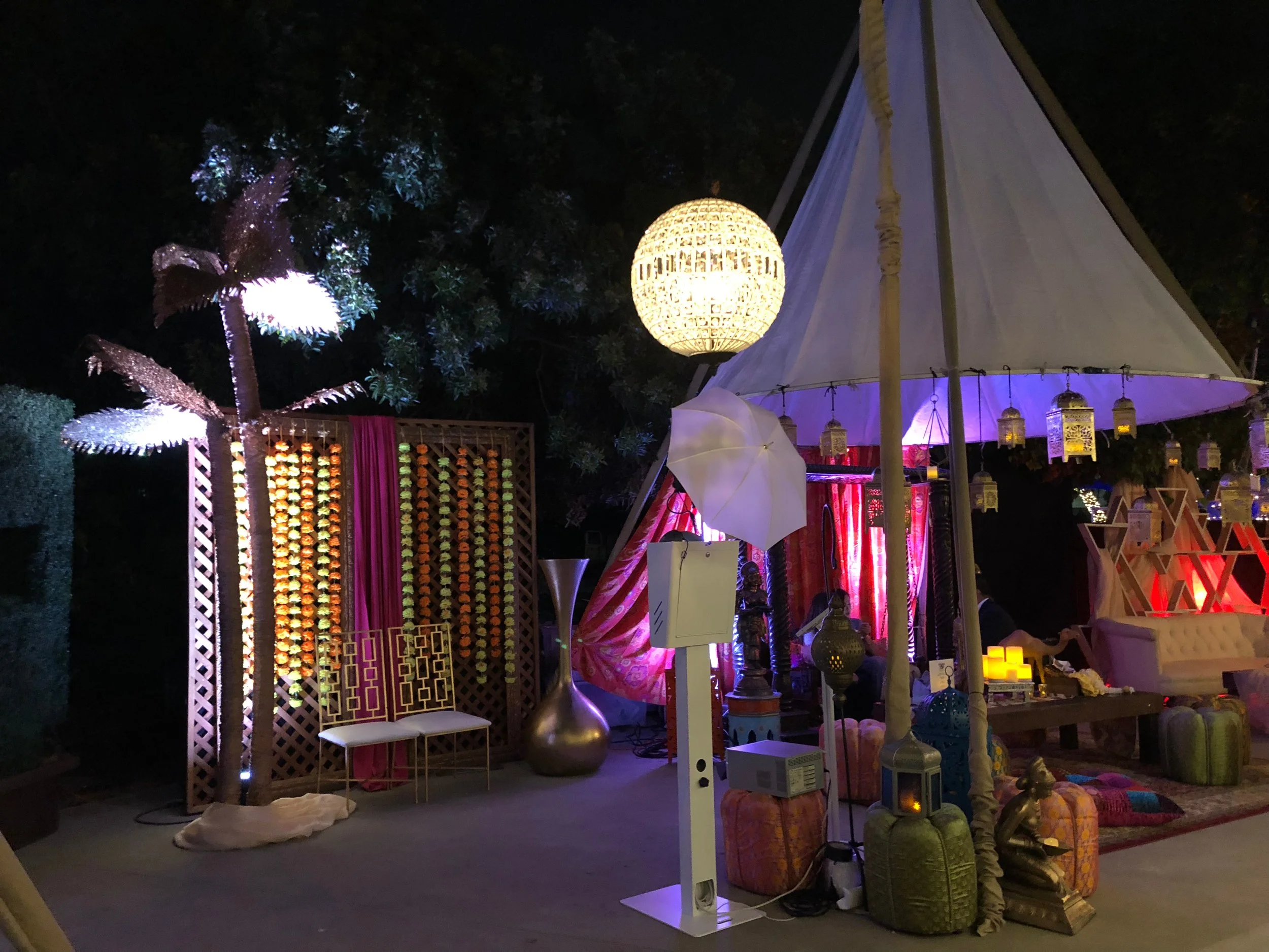 Decorative outdoor event setup with a large white tent, hanging lanterns, illuminated palm tree, and seating area with cushions and vintage accessories at night.