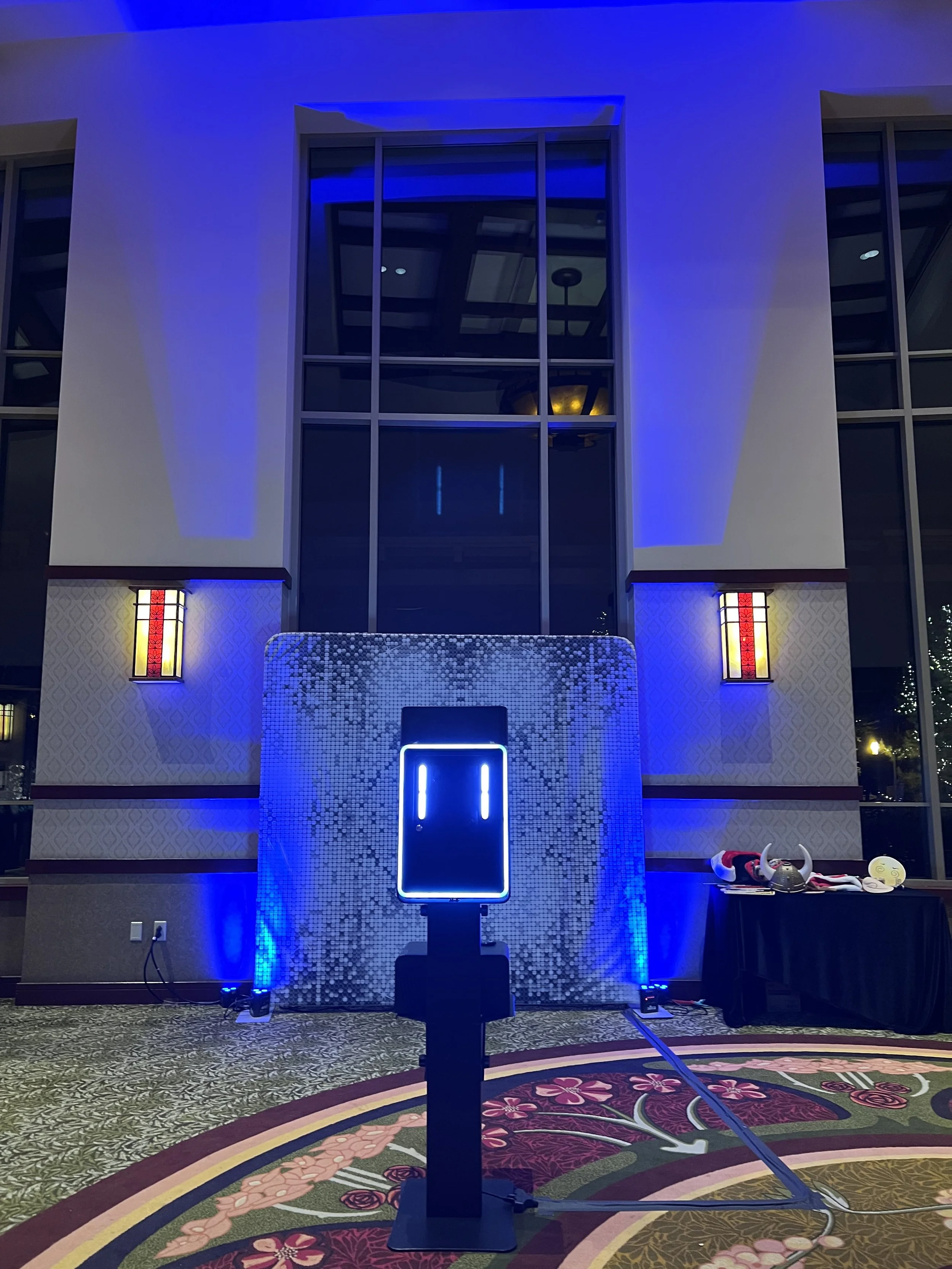 Indoor scene with a large windowed wall illuminated by blue lighting. There is a gaming or photo booth setup with a rectangular screen displaying two vertical lines, and decorations including tables with accessories, a patterned carpet, and wall scon