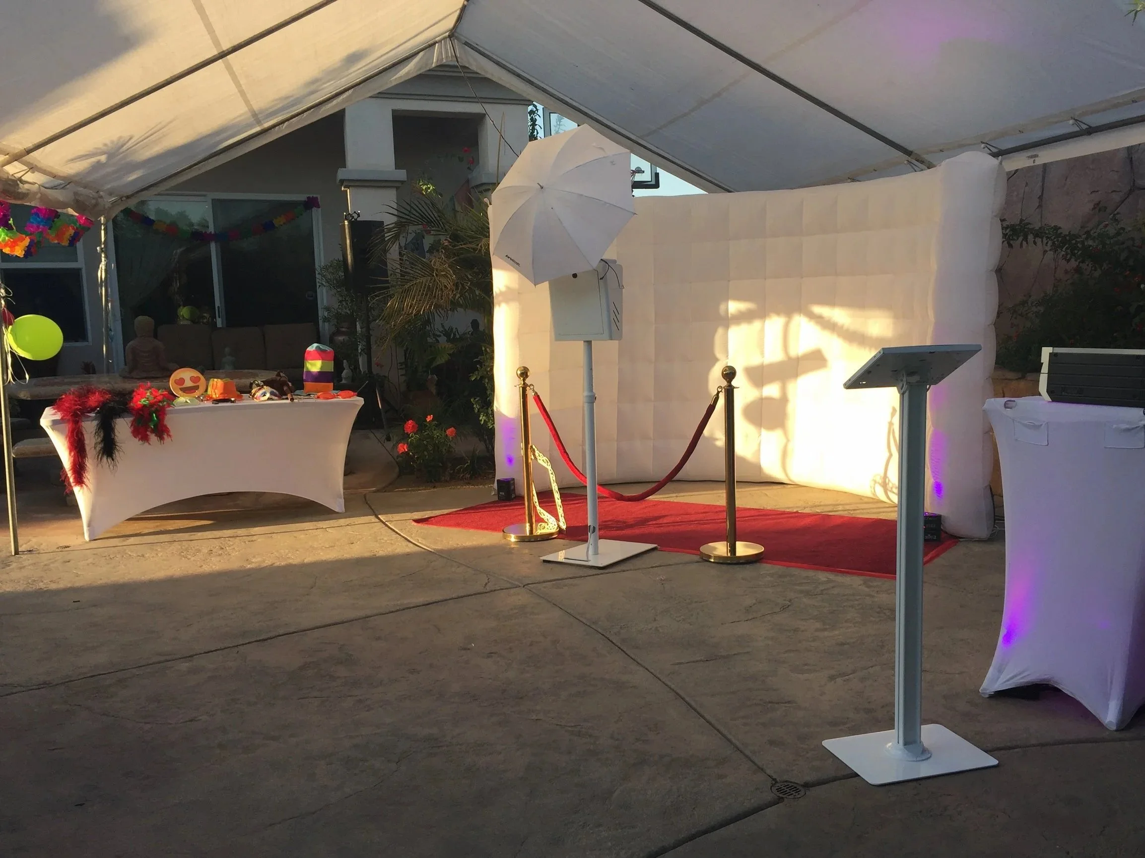 Party setup inside a white tent with a red carpet, velvet ropes, a selfie station with a backdrop, tables with decorations, and colorful party props.
