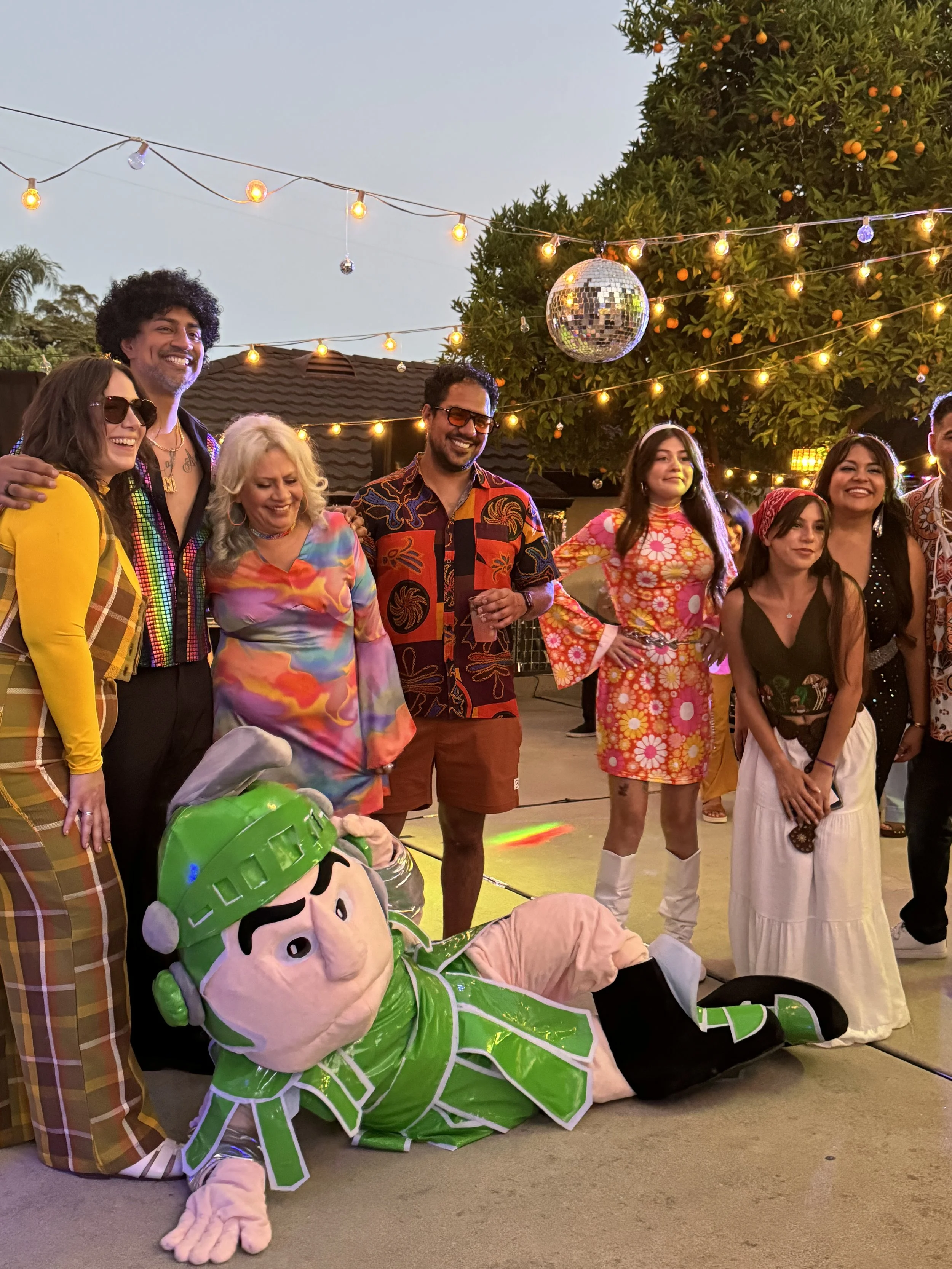 Group of people dressed in colorful 1960s-inspired outfits, standing outdoors under string lights and a disco ball, with a large orange tree behind them and a person in a Shrek costume lying on the ground in front.