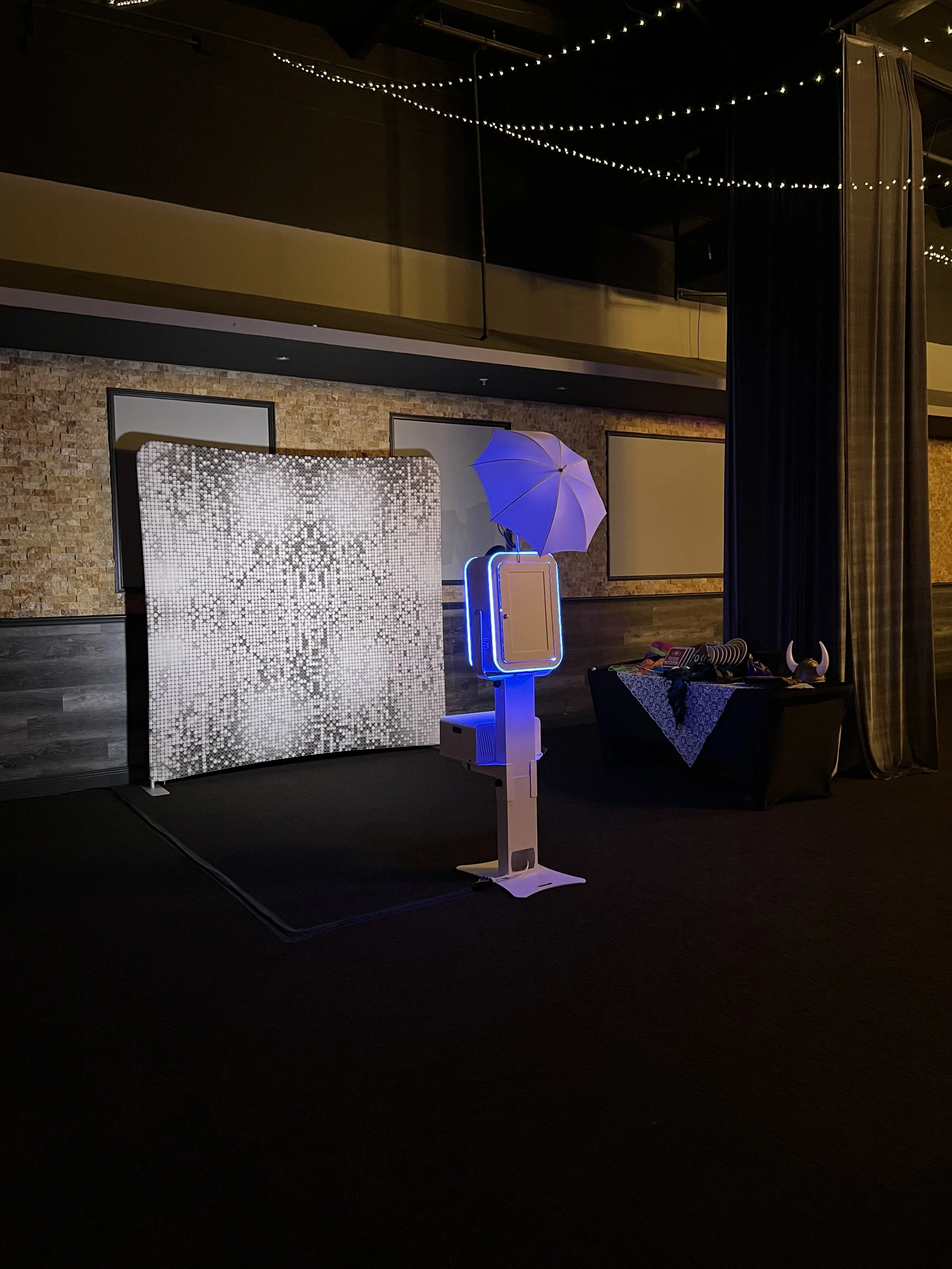 Photo of a photo booth setup with white umbrella, illuminated with blue lights, and a decorative backdrop in an indoor venue with string lights hanging from the ceiling.