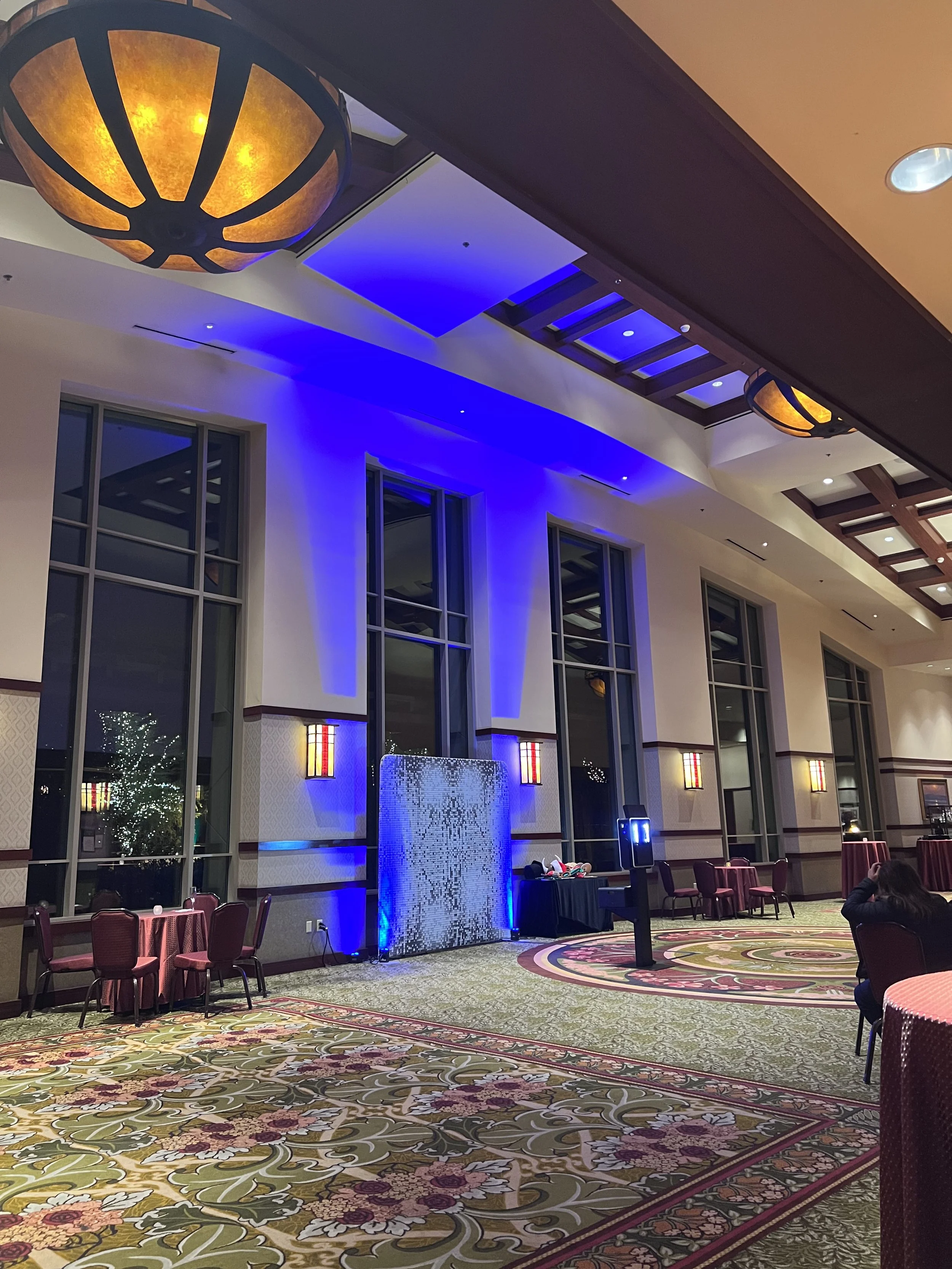 An event hall with high ceilings, tall windows, and a floral carpet. There are tables and chairs, blue lighting on the wall, and decorative lighting fixtures on the ceiling.