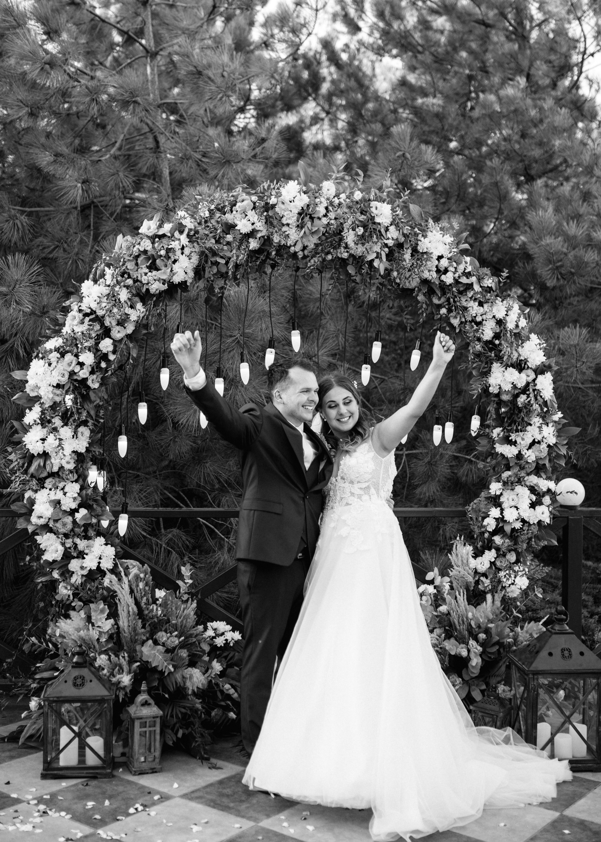 A newlywed couple celebrating under a floral arch with fairy lights in a garden setting, black and white photo.