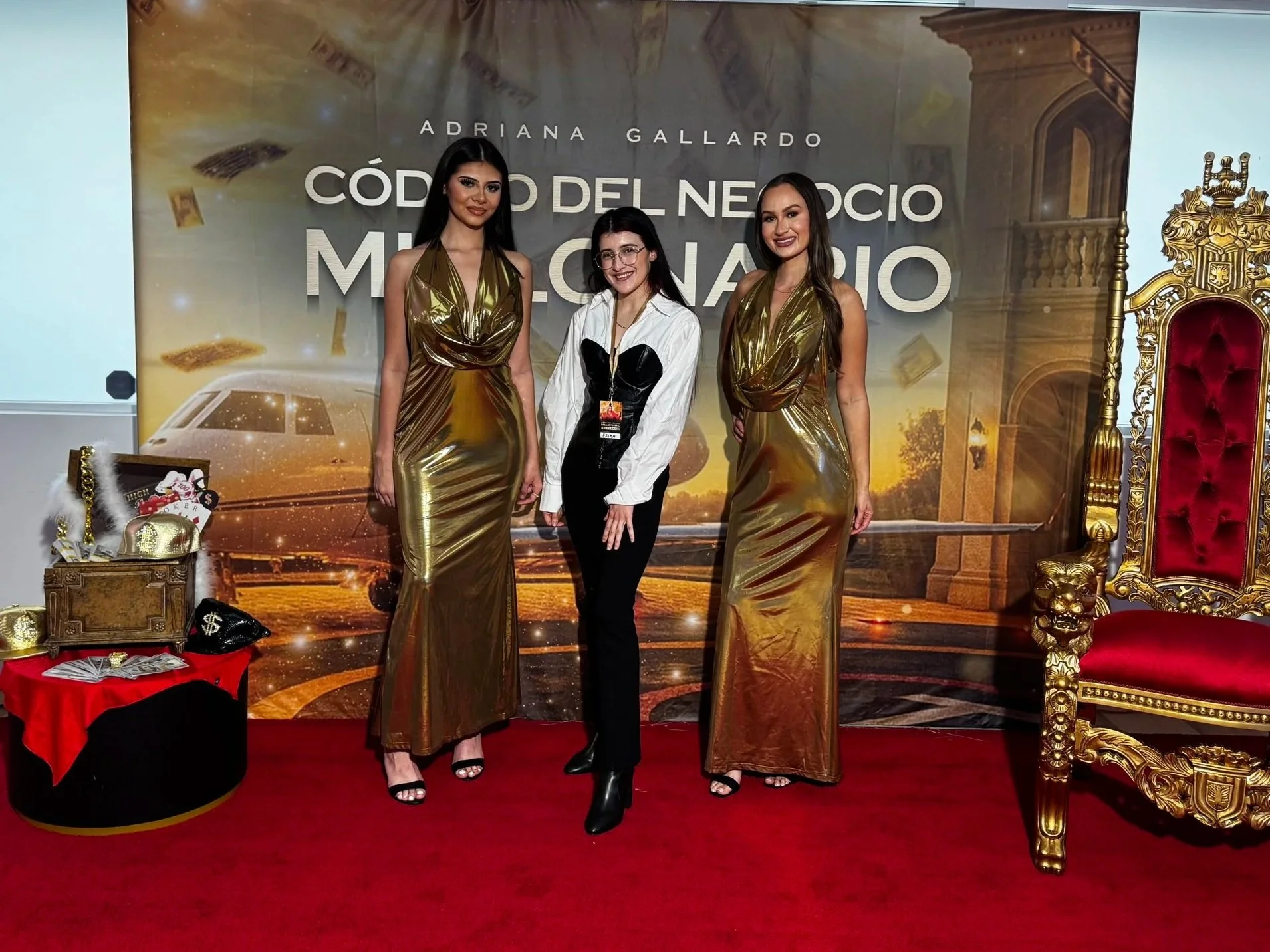 Three women standing on a red carpet in front of a promotional backdrop, with one woman in the center wearing a white shirt and black pants, and two women on each side wearing shiny gold dresses, during an event.