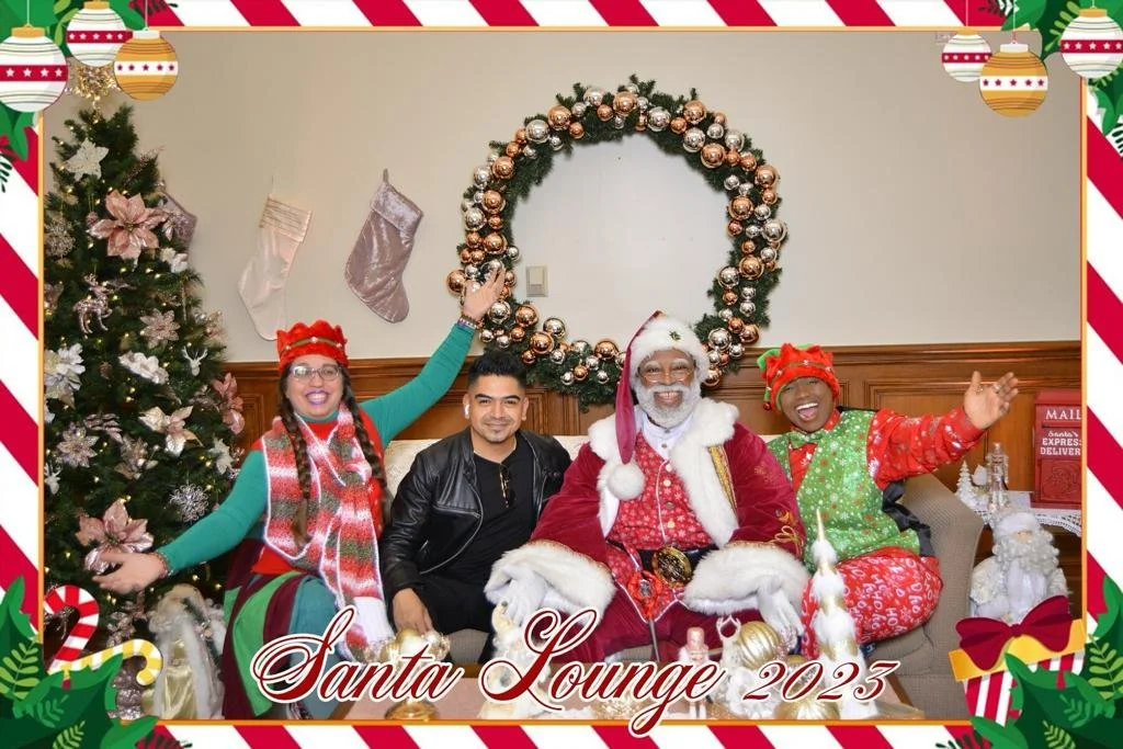 Group of four people, including Santa Claus, sitting in a Christmas-themed lounge with Christmas decorations and stockings hanging on the wall, celebrating 2023.