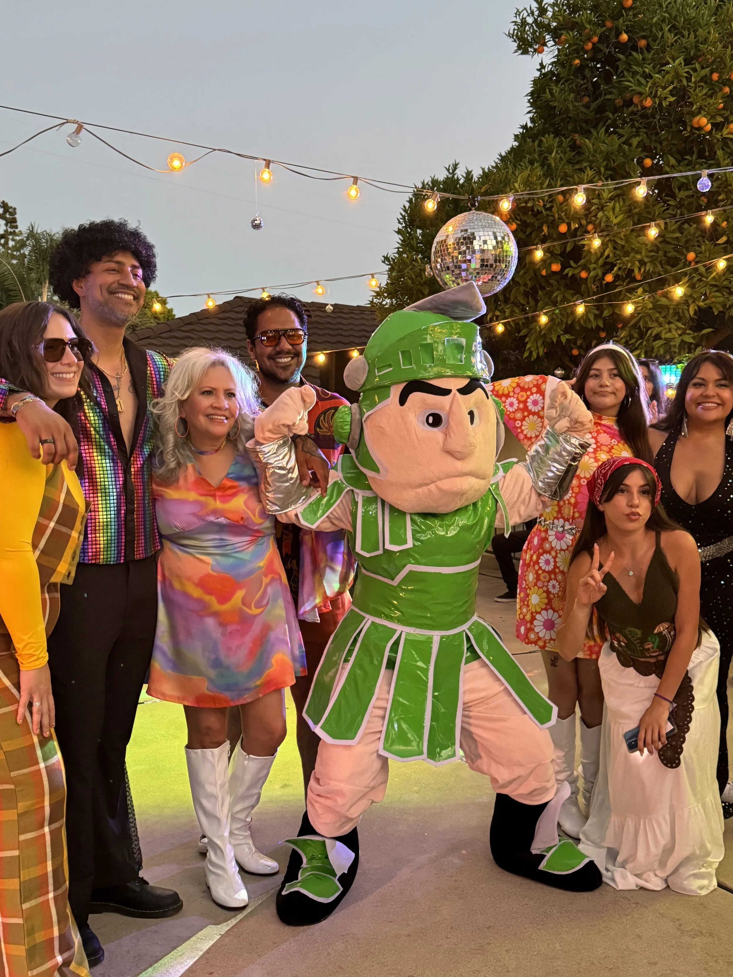 Group of people posing with a large mascot dressed as a character in a colorful costume, outdoors during evening with string lights and a tree with oranges in the background.