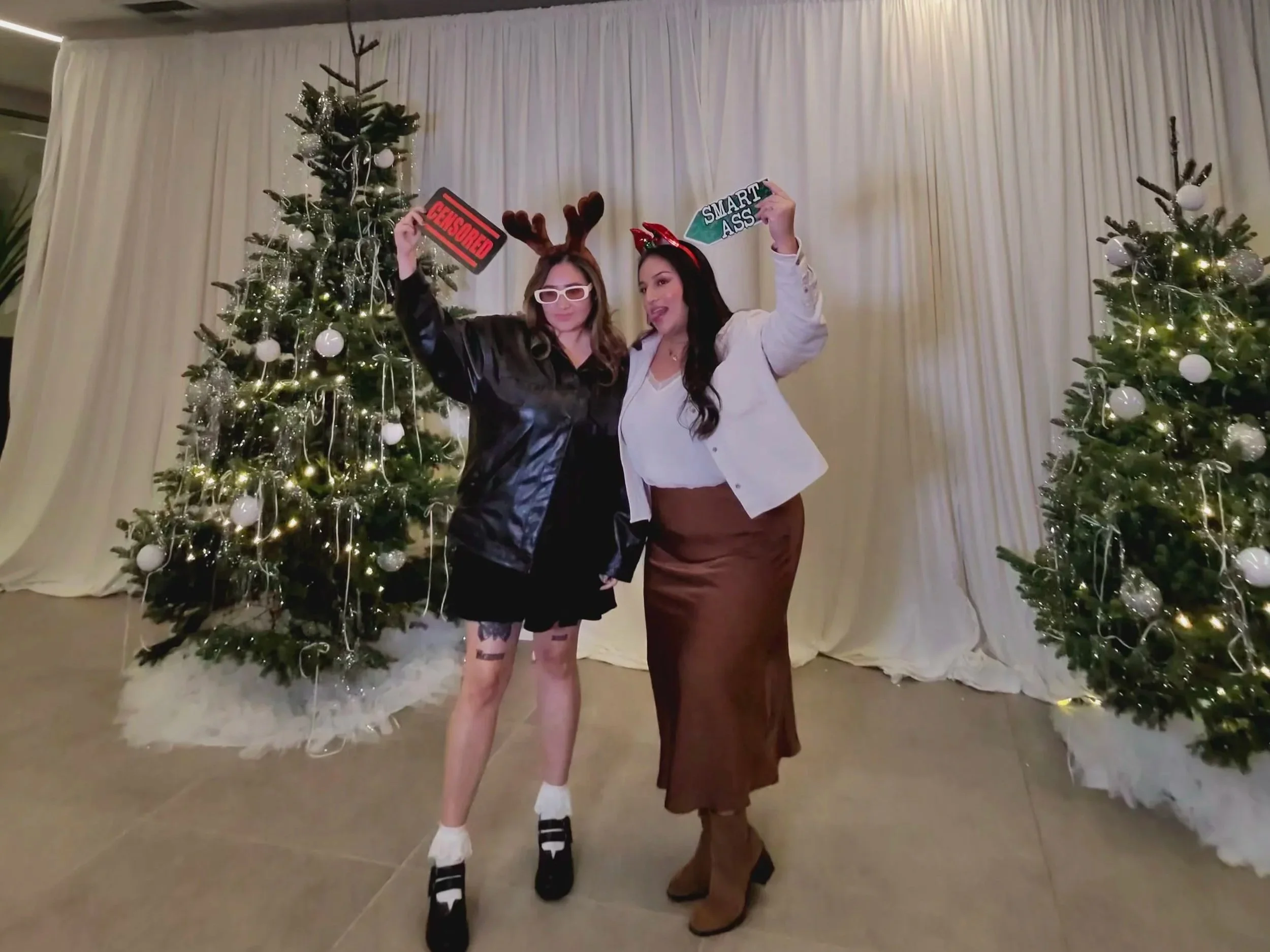 Two women in Christmas-themed clothing with holiday props, standing in front of decorated Christmas trees with white ornaments and lights, posing for a photo.
