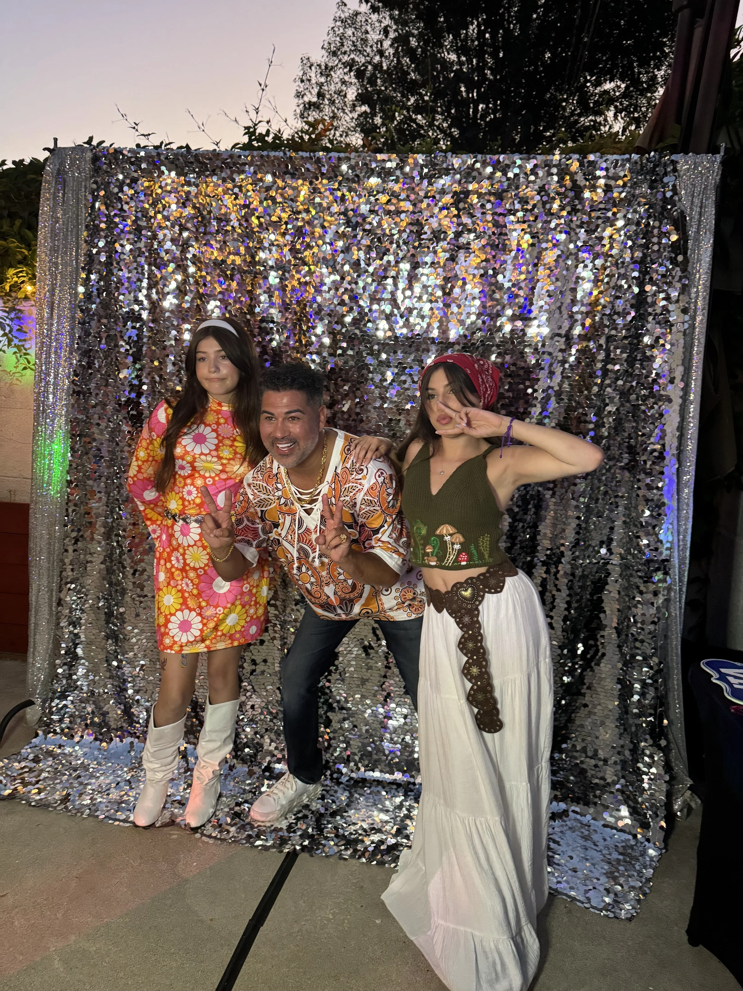 Three people posing in front of a sparkly silver backdrop at a party or celebration outdoors during the evening.