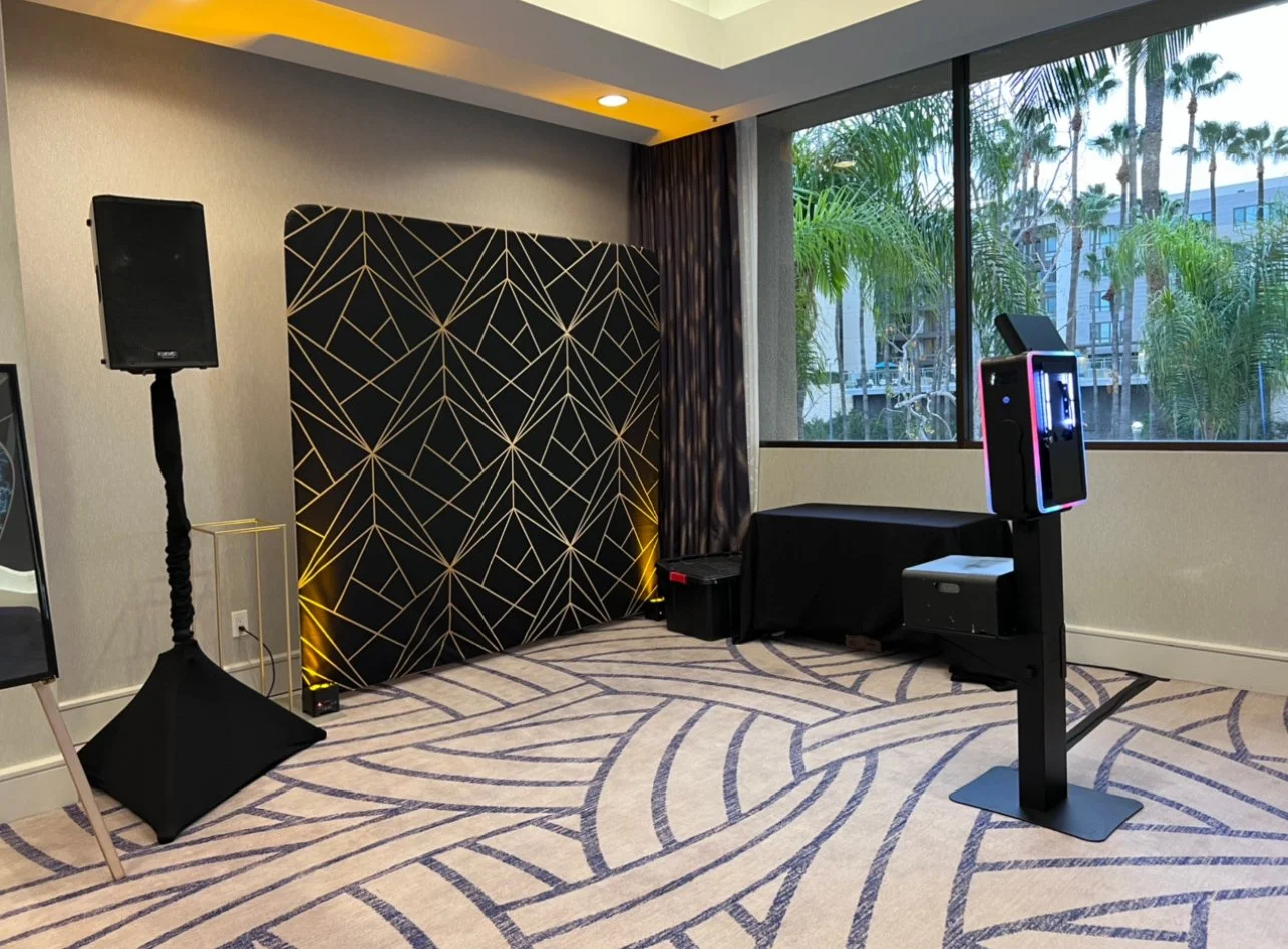 An indoor event space features a stage with a black and gold geometric backdrop, a speaker on a stand, and a photo booth with colorful neon lighting. There is a large window showing palm trees outside.
