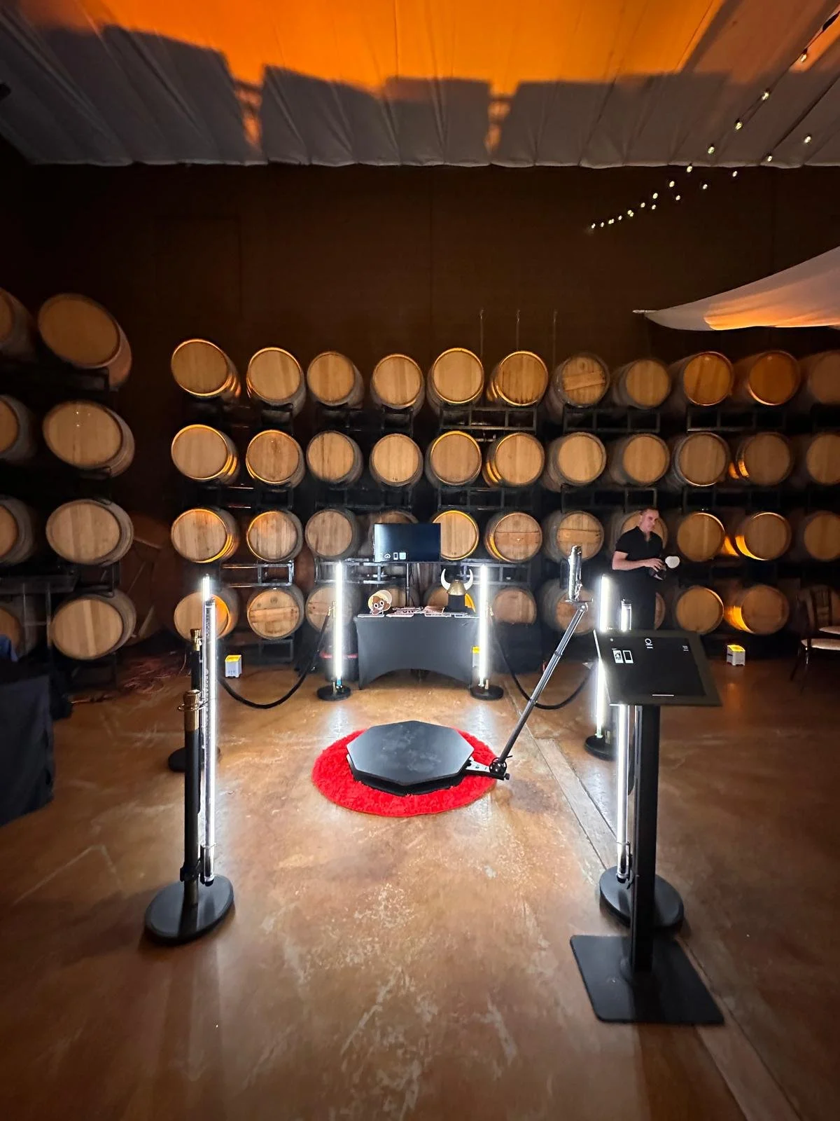 A room with wine barrels stacked on the wall, a red carpet in the foreground, a small display table, and a person standing on the right side.