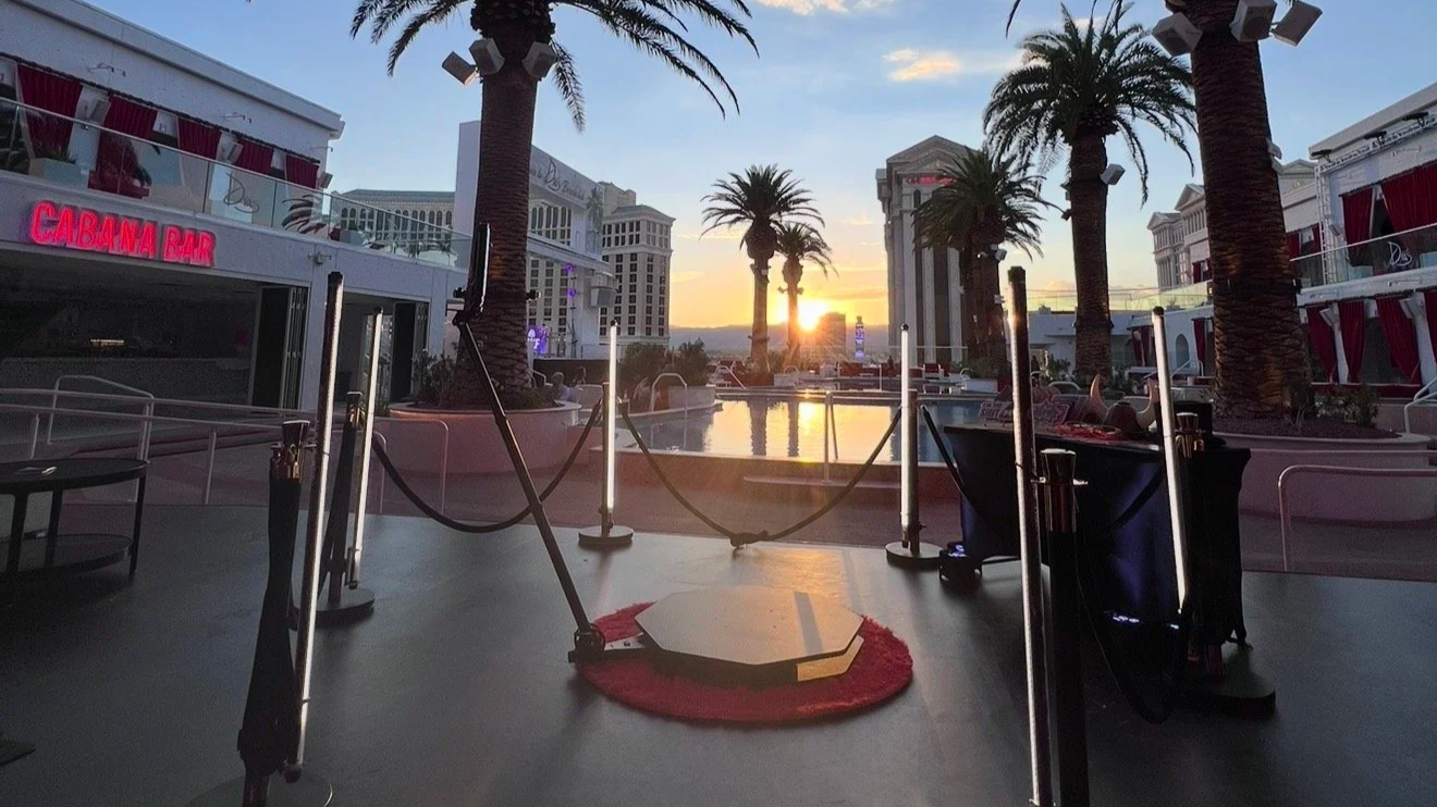 View of a poolside area at sunset with palm trees, white buildings, and a Las Vegas skyline in the background. There is a small stage or platform with a red carpet, stanchions, and ropes, and a neon sign that says 'CABANA BAR'.