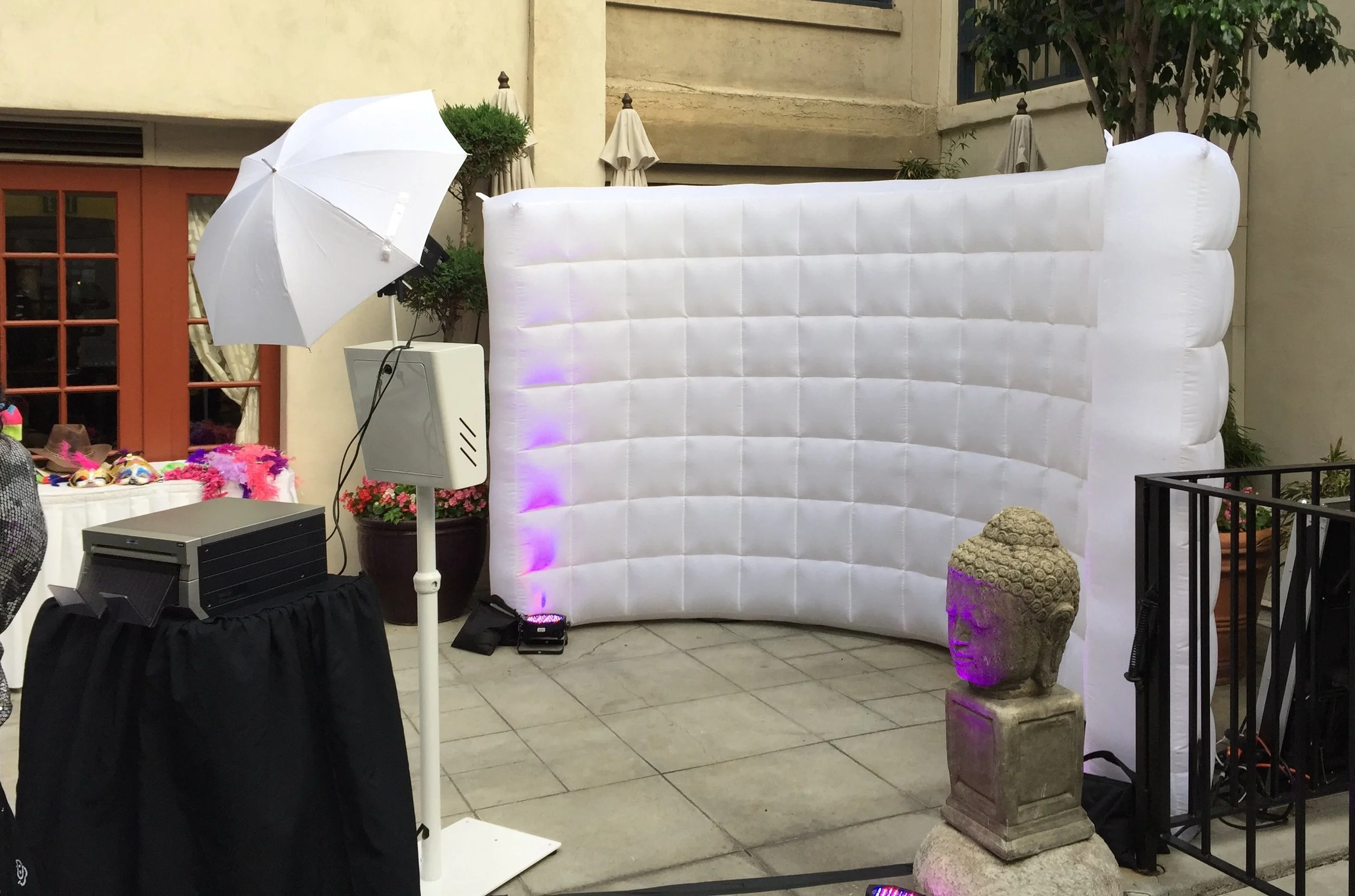 Inflatable photo booth with a white backdrop, purple lighting, a decorative stone Buddha statue in front, and a white umbrella light on a stand, set up outdoors on a tiled patio.