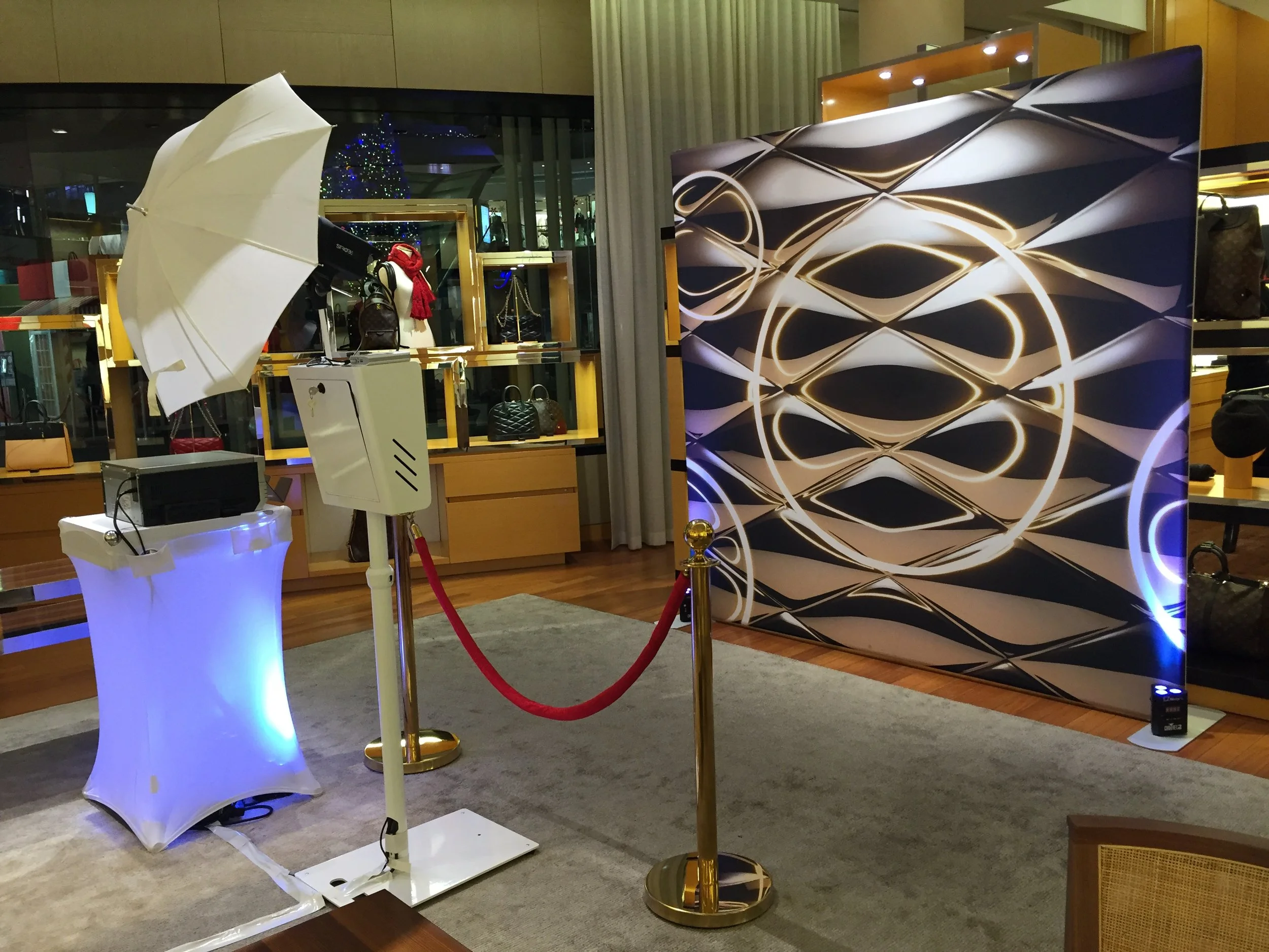Photo of a photo booth setup with a large abstract graphic background, a white umbrella, and a white stand with lighting equipment inside, situated on a gray carpet in an indoor shopping area with display cases and handbags.