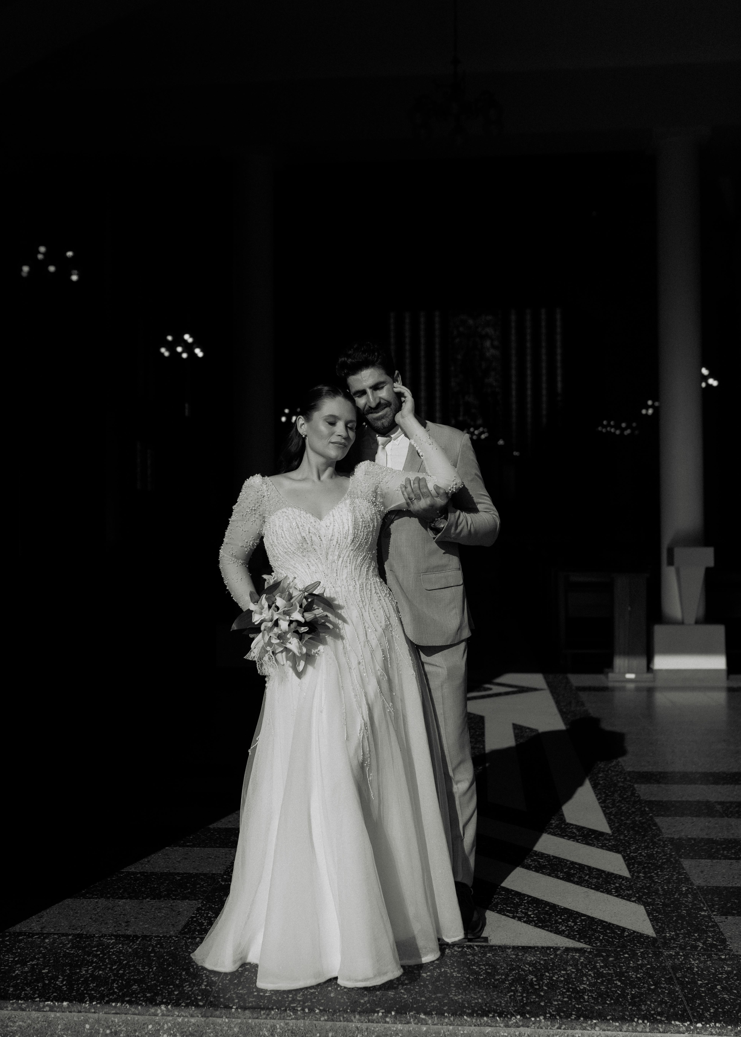Black and white photo of a bride and groom walking together, the bride in a wedding dress holding a bouquet, and the groom in a suit, inside a venue with columns and chandeliers.