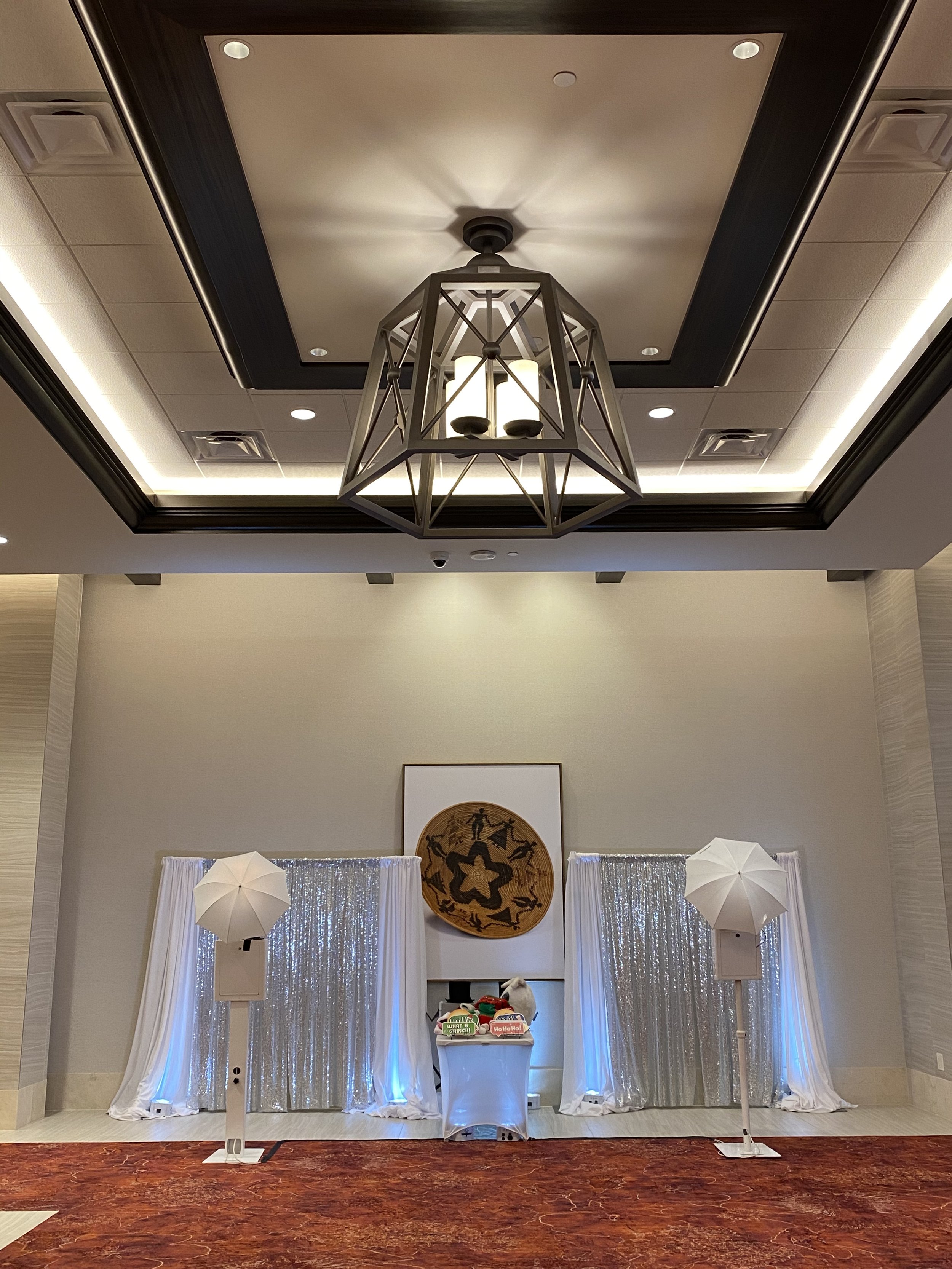 Indoor event setup with a backdrop of silver curtains and a white table in front of it, flanked by two white umbrellas on stands. The table holds Christmas-themed decorations, with a framed artwork with tribal or folk art style above the setup. Overhead, a modern geometric chandelier hangs from a ceiling with recessed lighting and decorative trim.