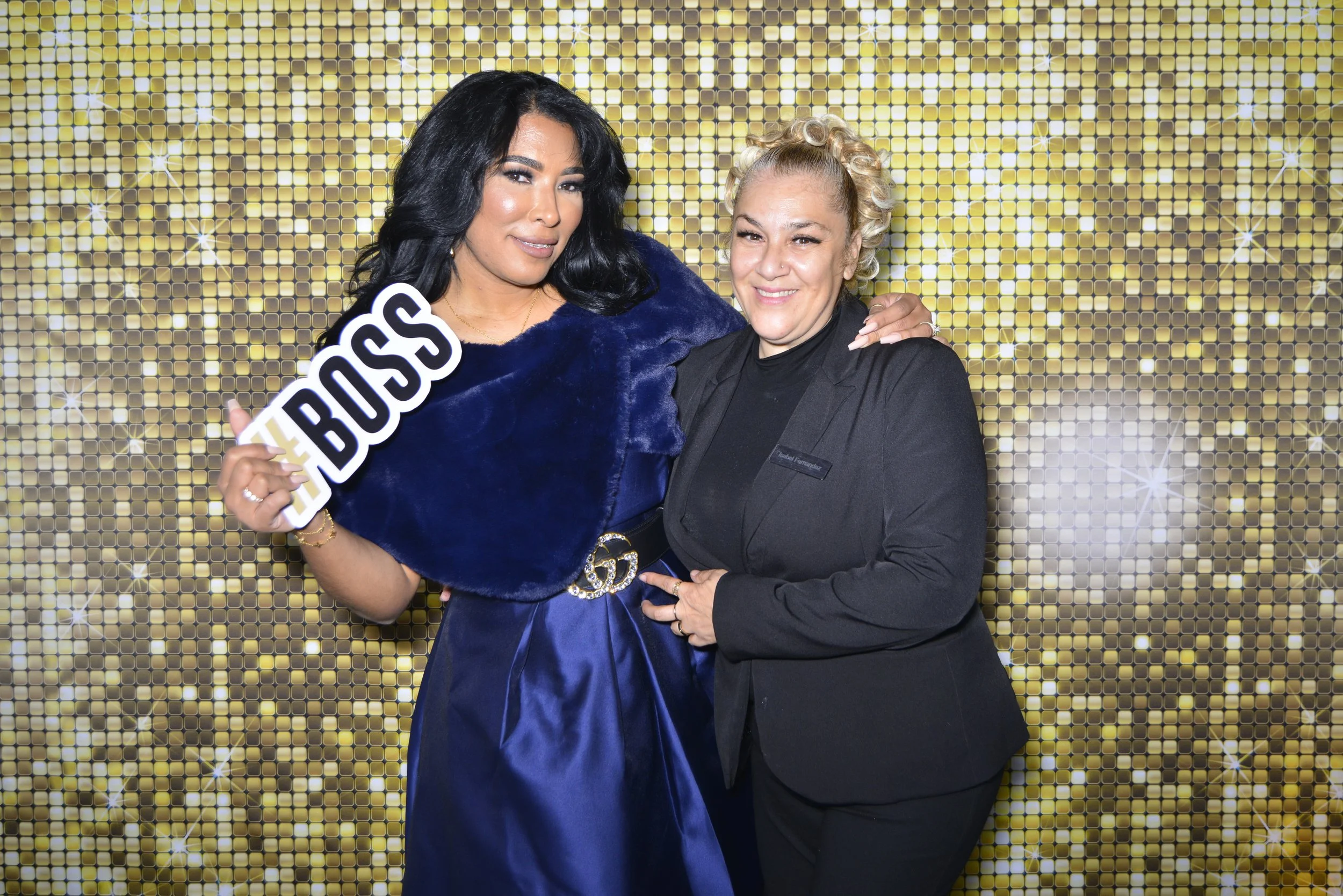 Two women posing together in front of a gold sequin backdrop. One woman is holding a sign that reads '#BOSS' and the other has her arm around her. The woman on the left is dressed in a royal blue outfit with a black belt, while the woman on the right