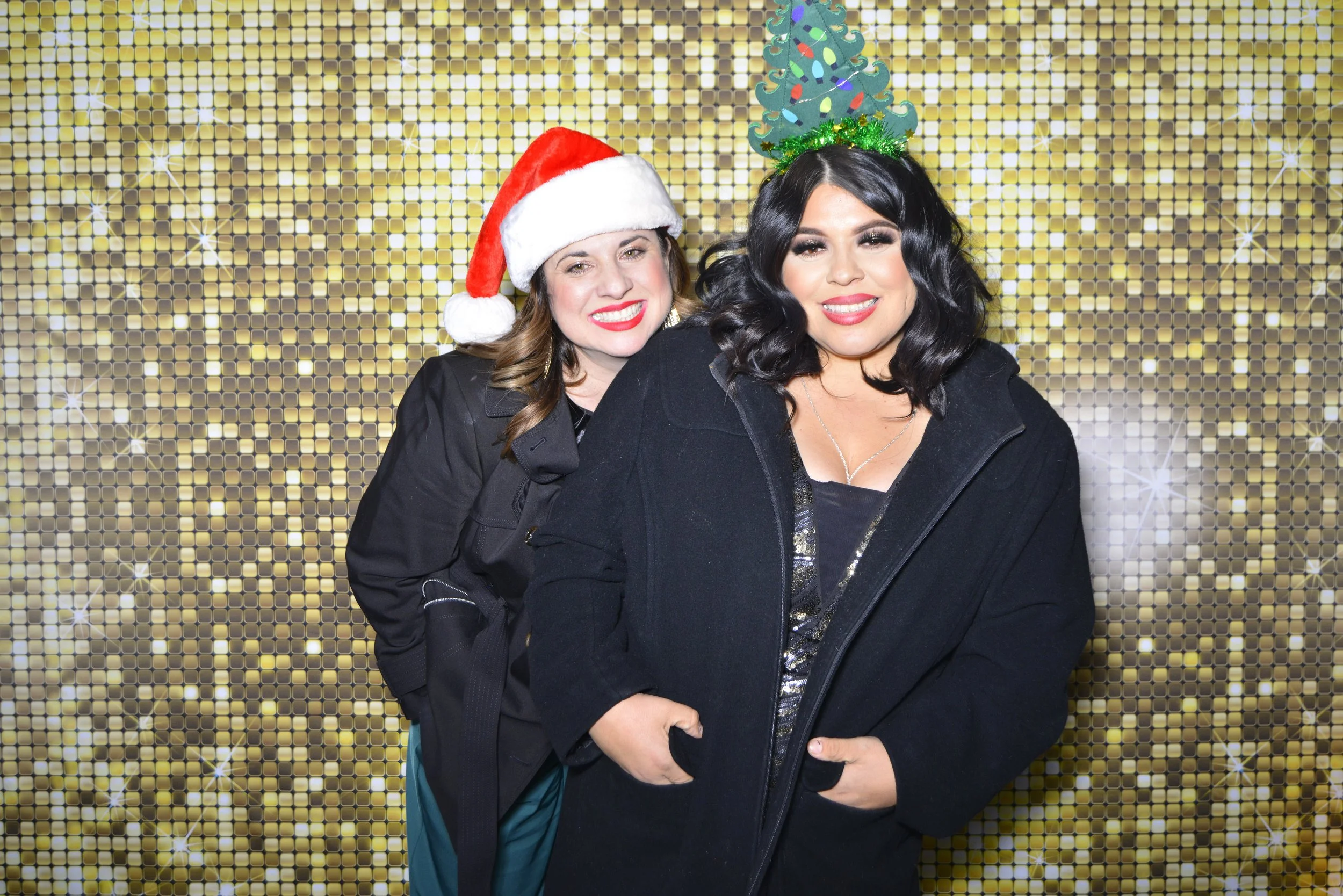 Two women smiling in front of a gold sparkly backdrop. One woman is wearing a Santa hat, and the other is wearing a Christmas tree headband.