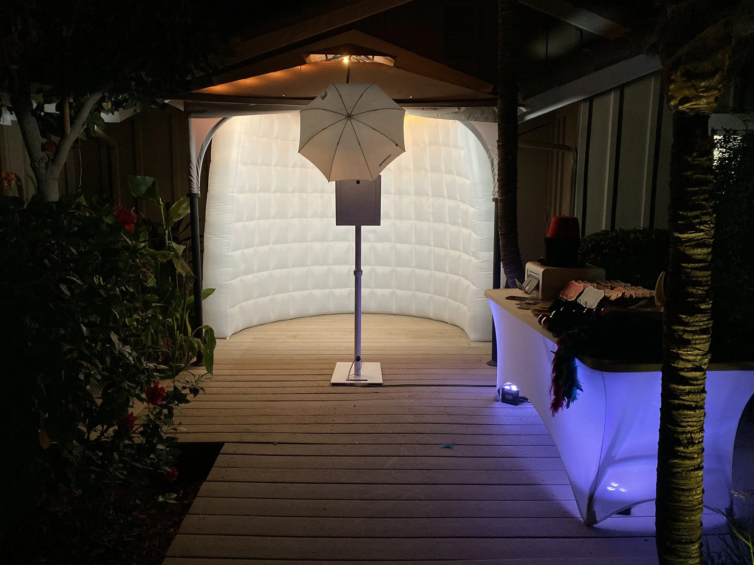 An outdoor photo of a small bar setup with a white tablecloth, a computer, and a shelf with supplies. There is a white umbrella and a white inflatable backdrop with ambient lighting, surrounded by plants and trees at night.