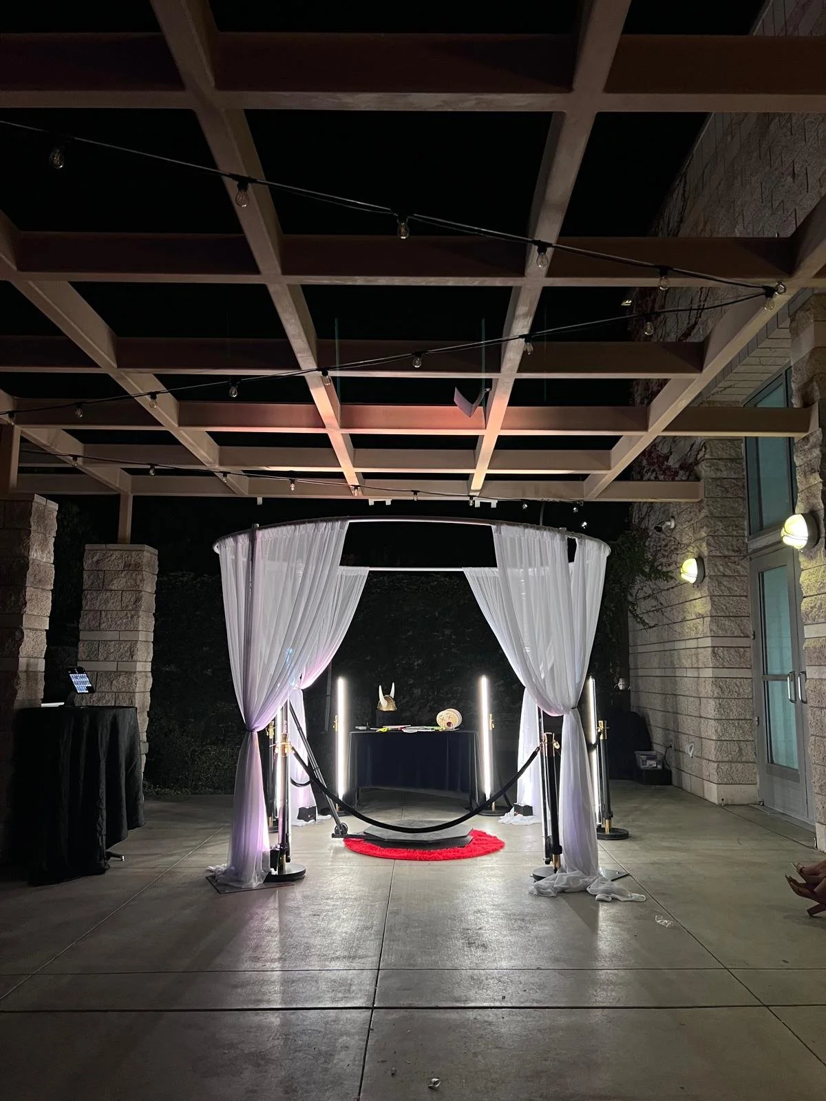 Photo of an outdoor wedding or party setup with a semi-circular white curtain canopy, a table with decorations, and a red circular rug on the ground. String lights hang above, and stone walls surround the area.