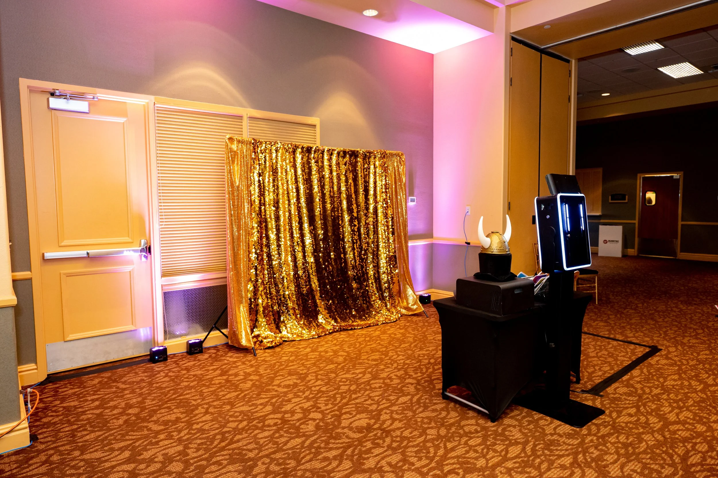 Photo of a photo booth setup with a gold sequin curtain backdrop and a black stand with a horned helmet, photo booth equipment, and a mirror photo booth in a decorated event space with pink and purple lighting.
