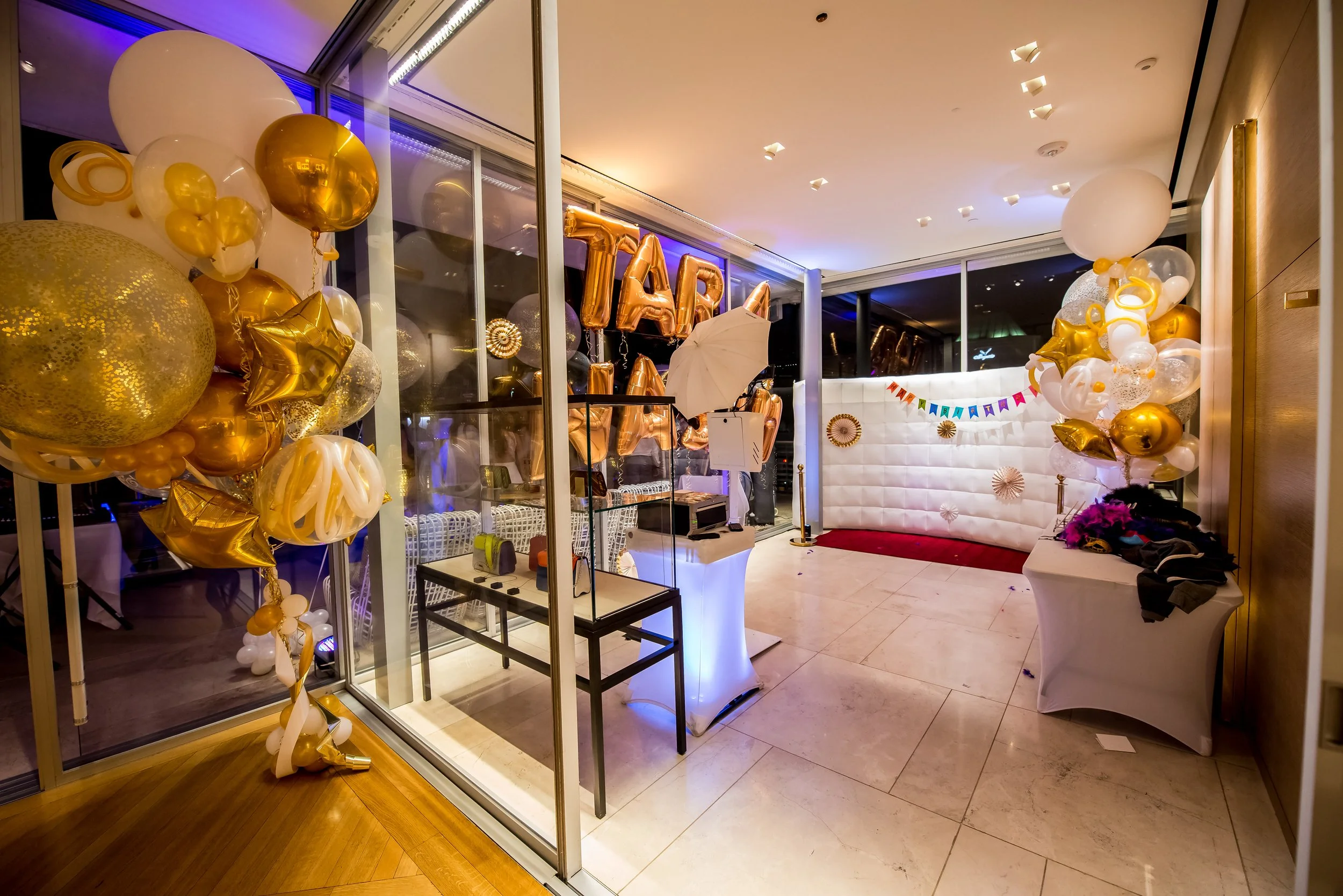 Party celebration area with gold and white balloons, a 'TARA' balloon banner, and a 'HAPPY BIRTHDAY' banner, decorated with gold and white accents, set up inside a room with glass walls and a tiled floor.