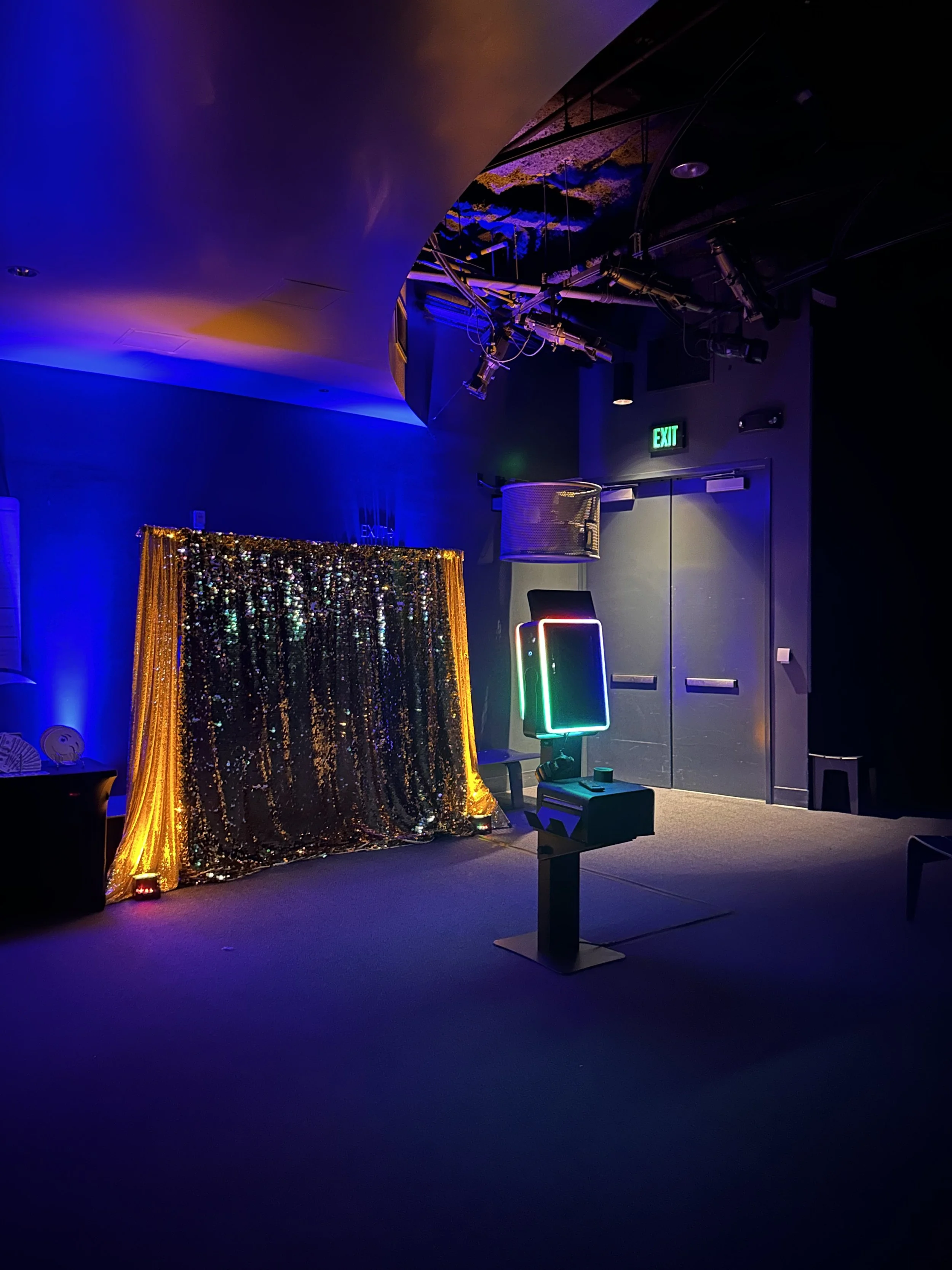 A photo of a dark room with colorful lighting, a glittery gold backdrop, and a photo booth with glowing neon lights.