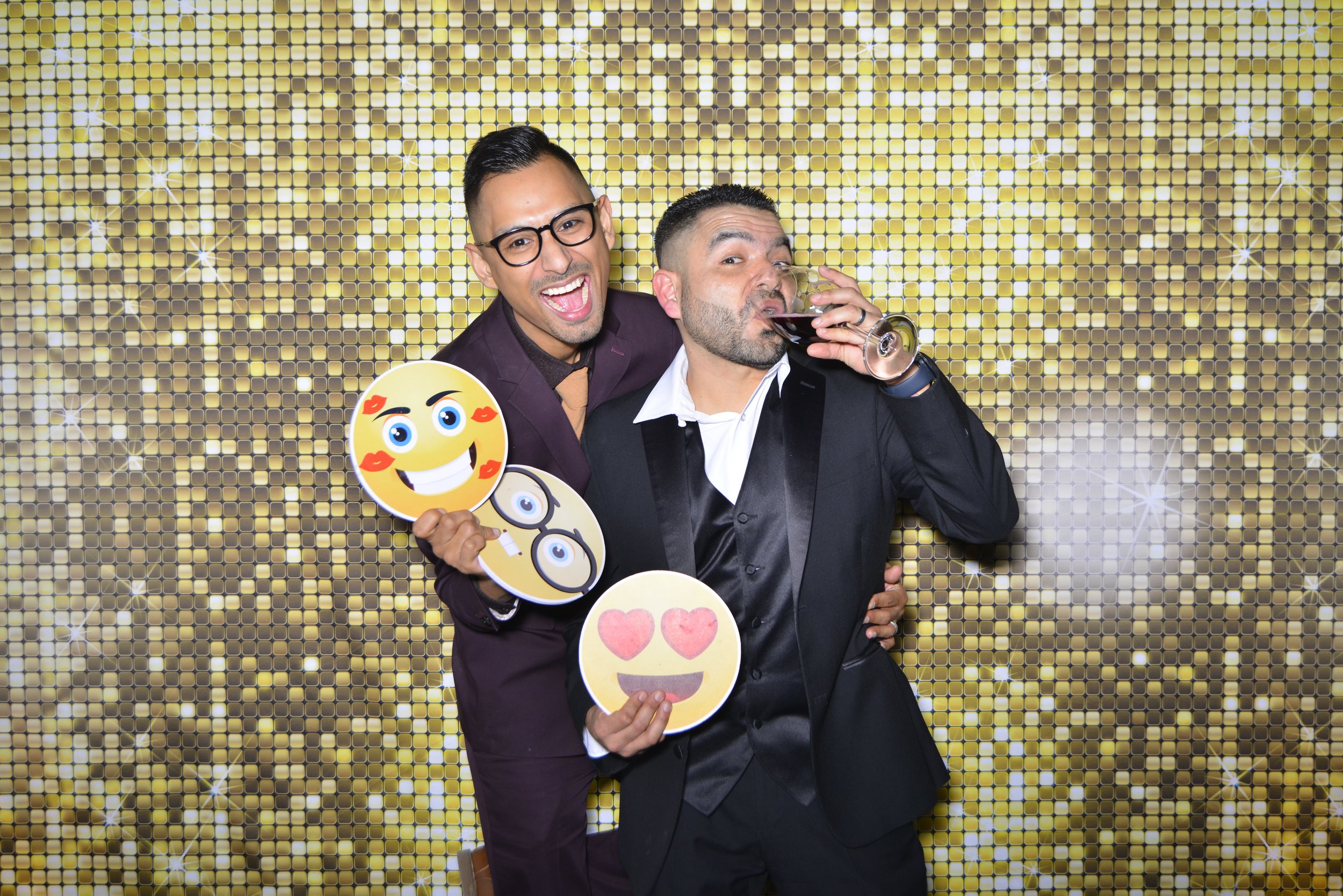 Two men at a celebration or party standing in front of a gold sequin backdrop. One man is smiling broadly with glasses, wearing a dark suit and holding emoji signs. The other man is drinking from a glass of red wine or cocktail, with a smile, wearing