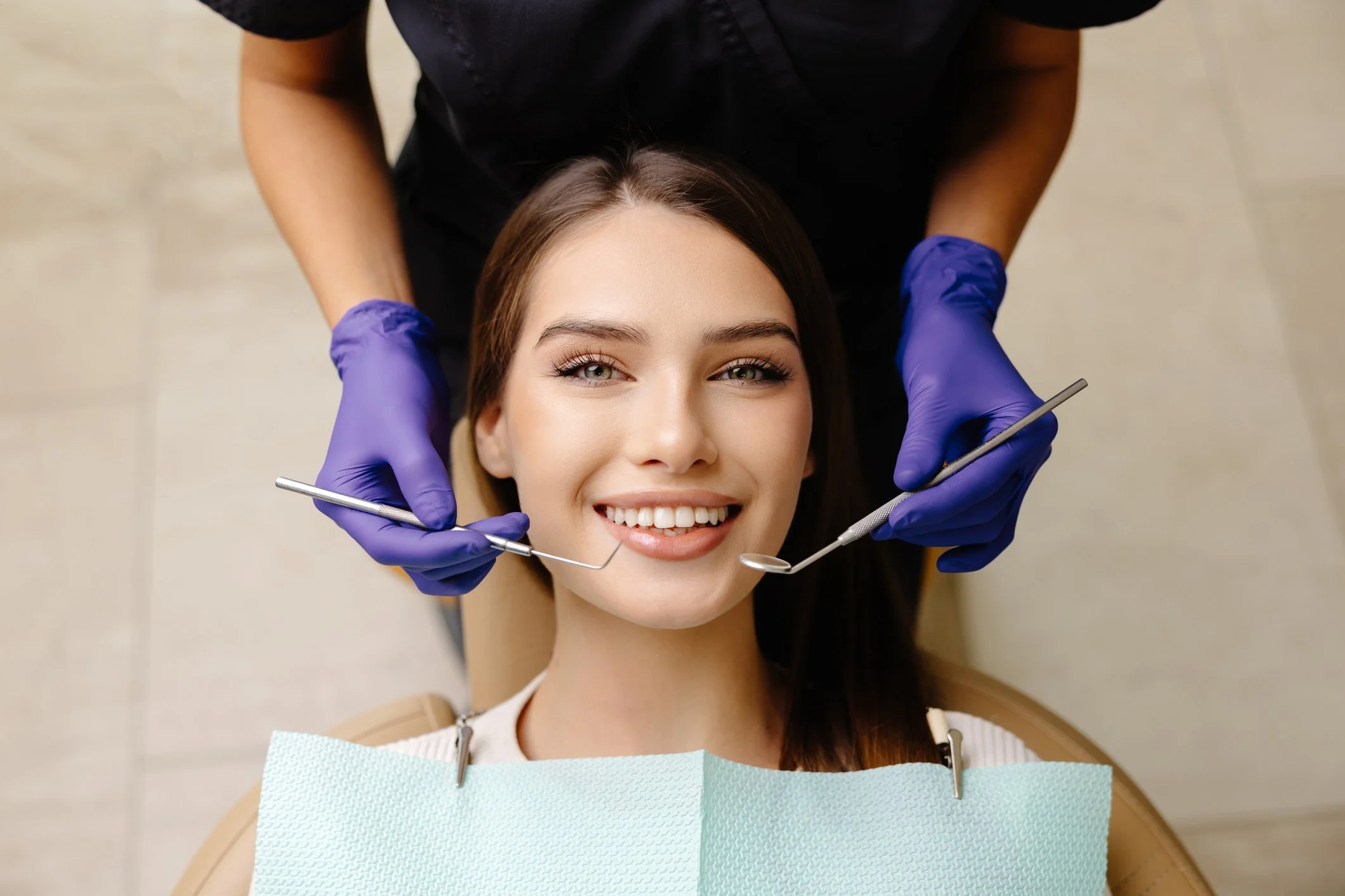 A woman with long dark hair and light skin smiling at the camera, lying back with her head on a dental chair. Two hands wearing blue gloves are holding dental tools near her mouth, and a dental bib is draped over her chest.