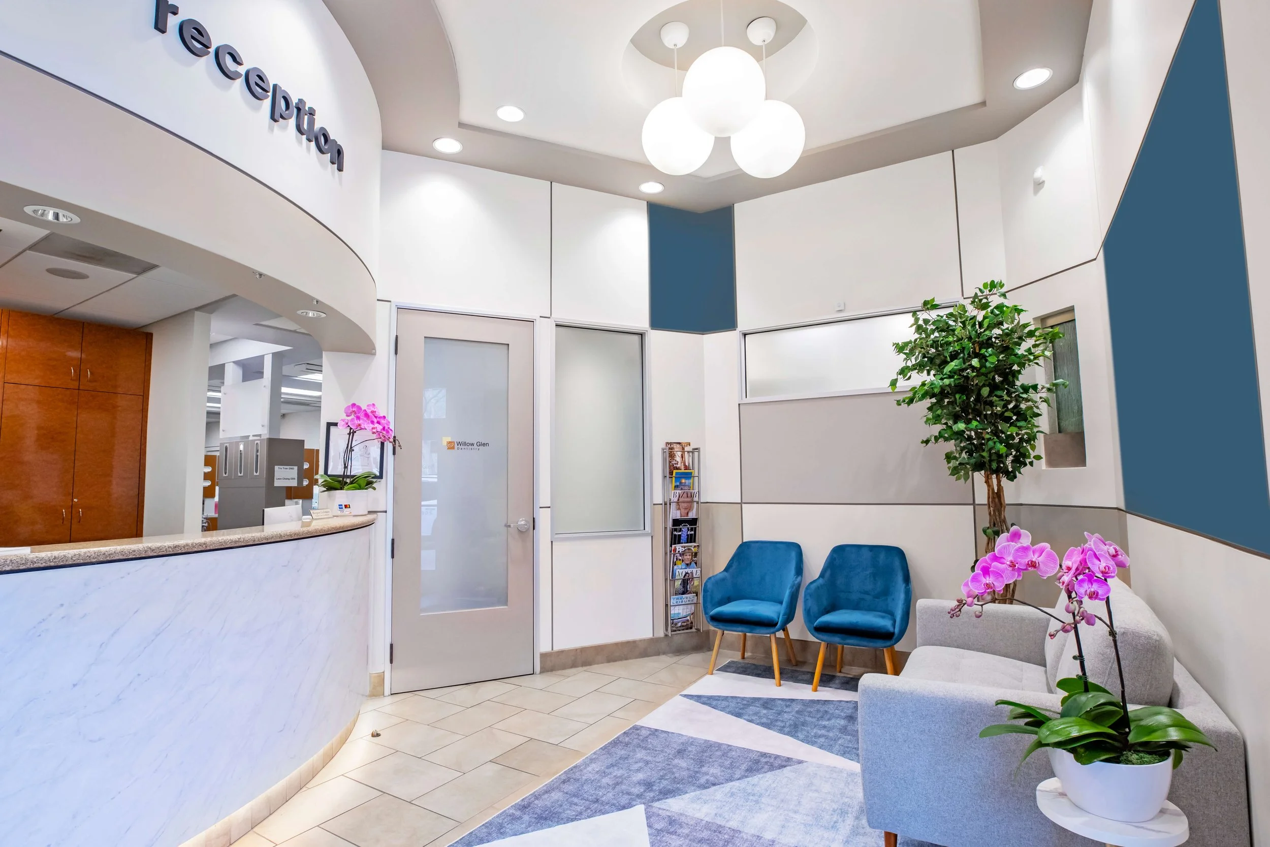 A modern , clean, comfortable, dental office reception area with blue chairs, potted plants, a white marble reception desk, and a waiting magazine stand. The walls are white with blue accents, and there are decorative lighting fixtures on the ceiling