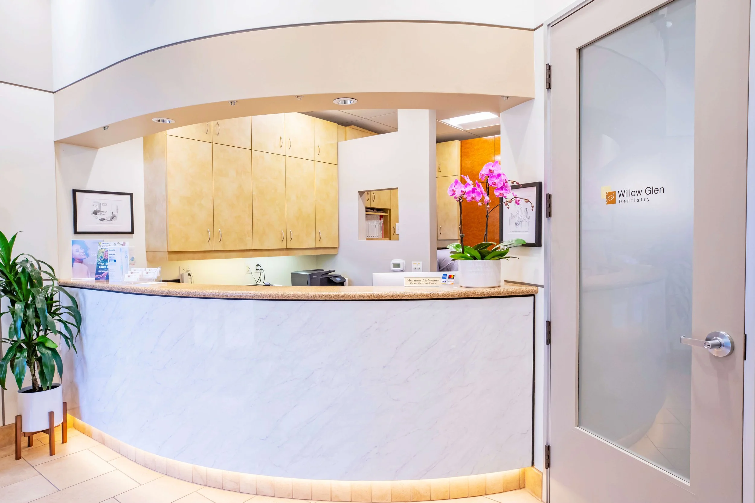 Reception desk with a pink orchid plant in a white pot, a computer, and pamphlets, with wooden cabinets behind the desk and framed artwork on the wall. The door to the right has the sign 'Willow Glen Dentistry'.
