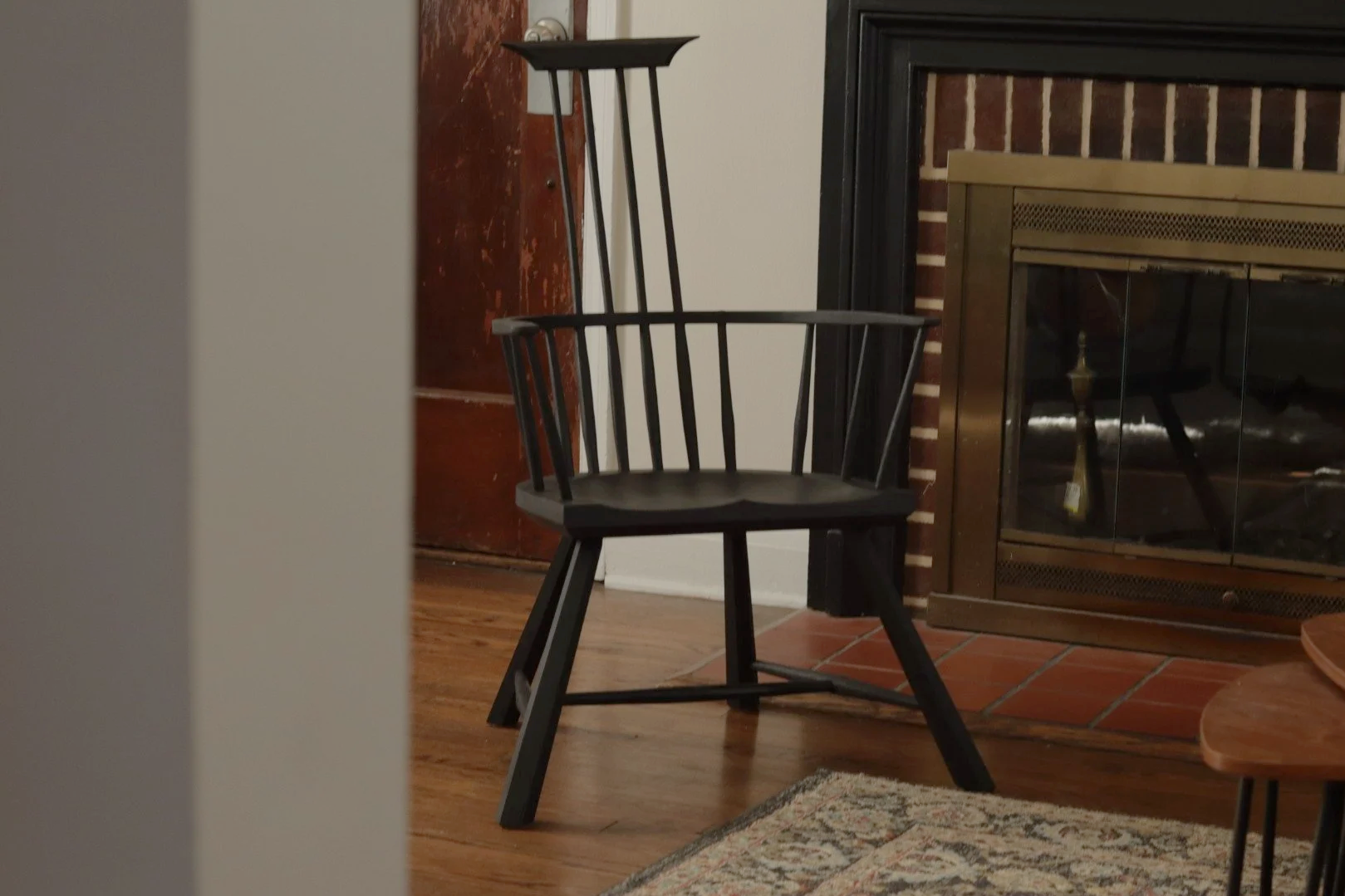 A black wooden chair with a spindle backrest in front of a fireplace with a brick surround and glass doors, wooden flooring, a colorful patterned rug, a white wall with a dark wooden door behind the chair, and part of a wooden table on the right side