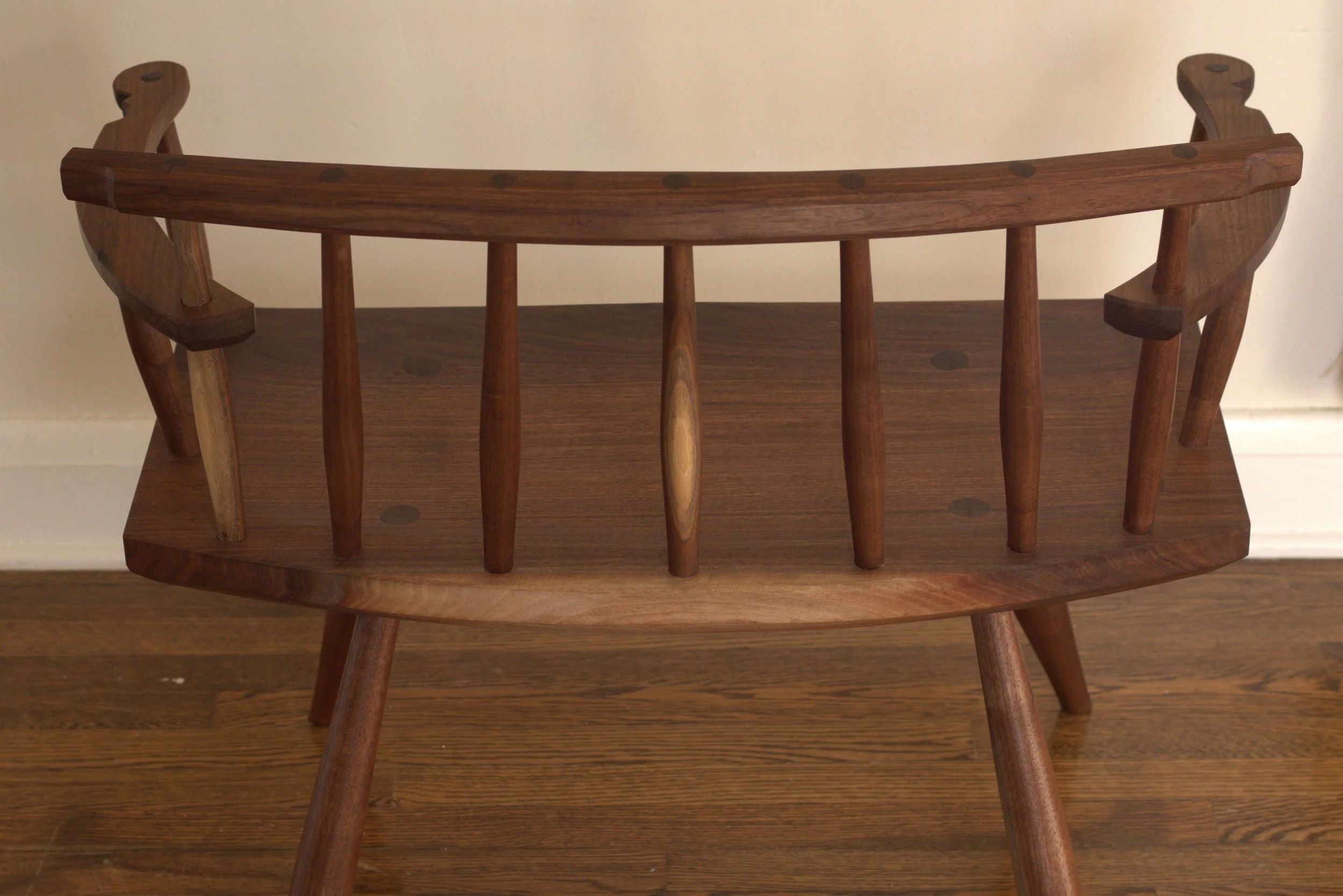 A modern styled wide low back chair made from walnut, with a backrest, spindles, and angled legs with rake and splay, placed against a white wall on a wooden floor.