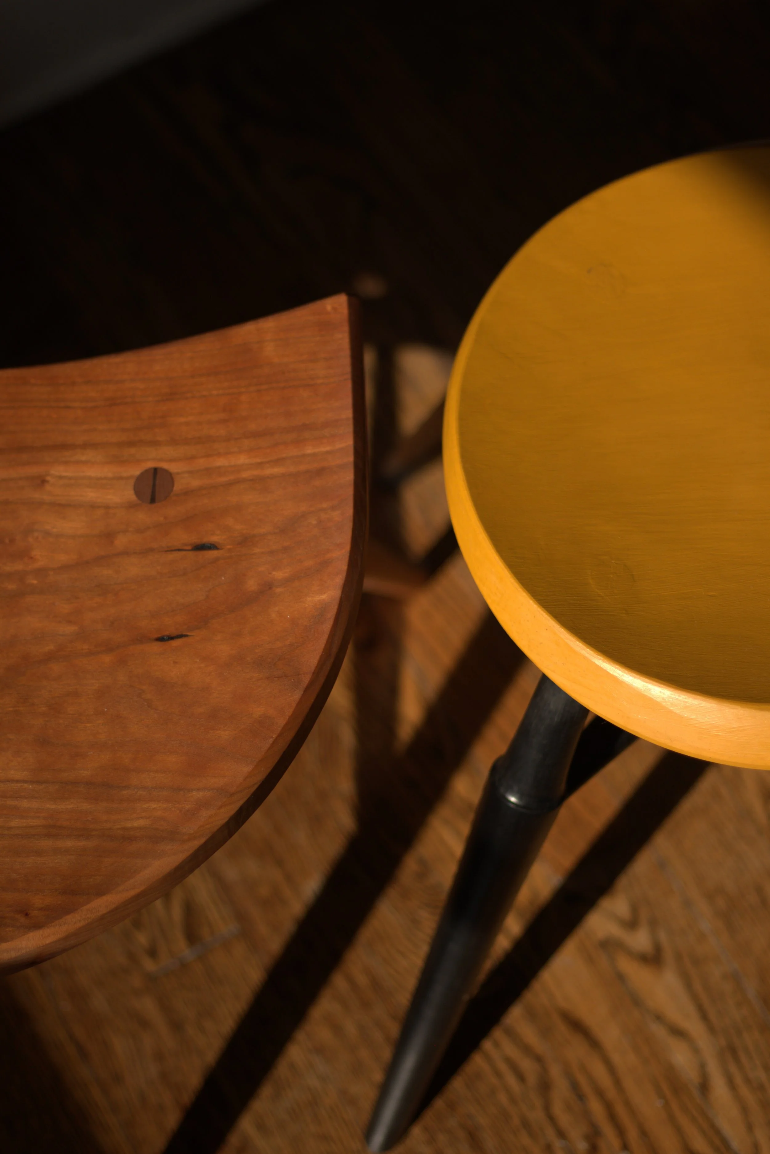 Close-up of wooden furniture, showing the edge of a cherry wood seat with visible through-tenon, and part of a yellow wooden stool with black legs, casting shadows on a wooden floor.