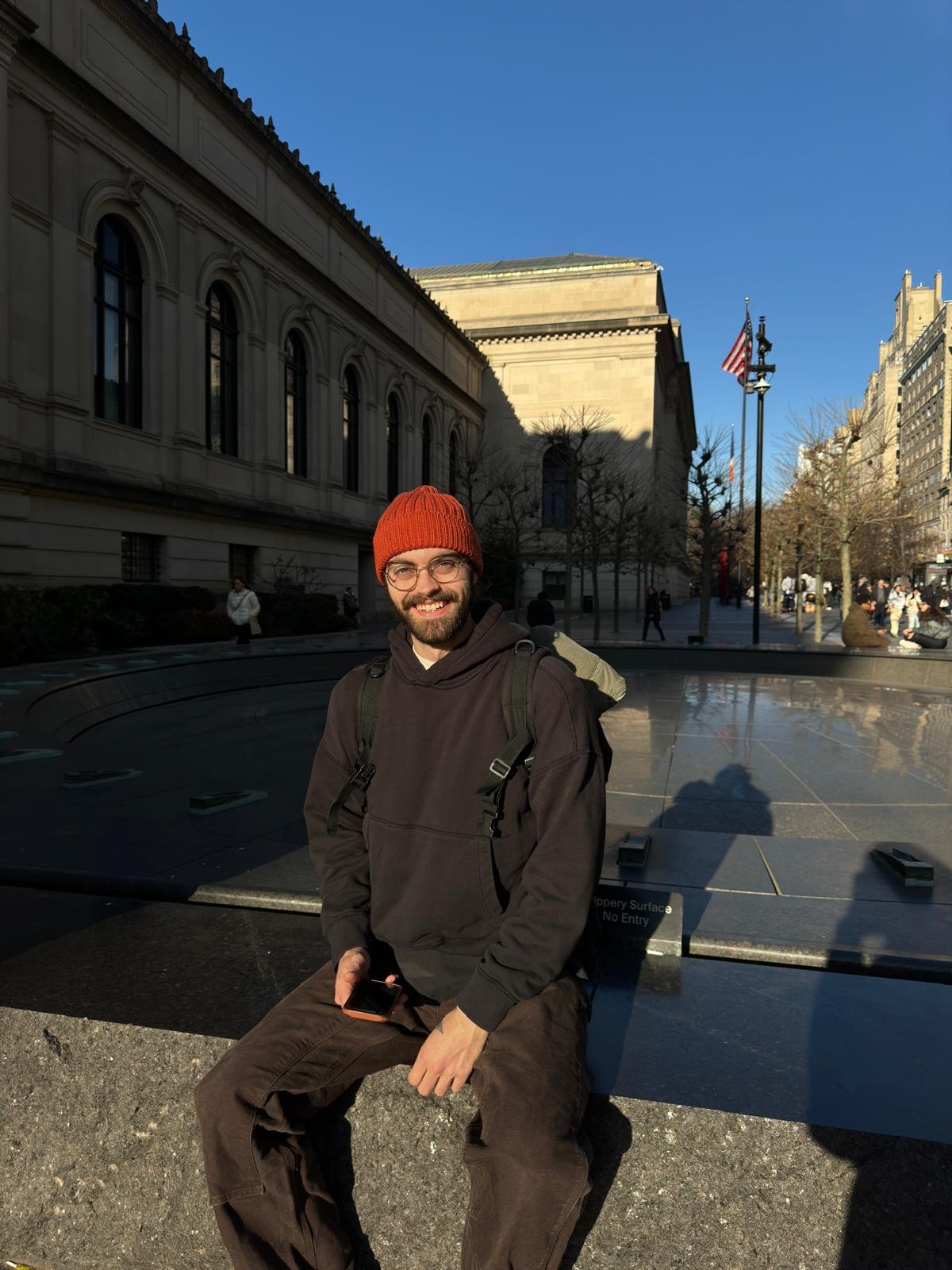 A man with a beard, glasses, and a red beanie sitting on a stone ledge in front of a fountain outside a large building with classical architecture, on a sunny day with a clear blue sky.