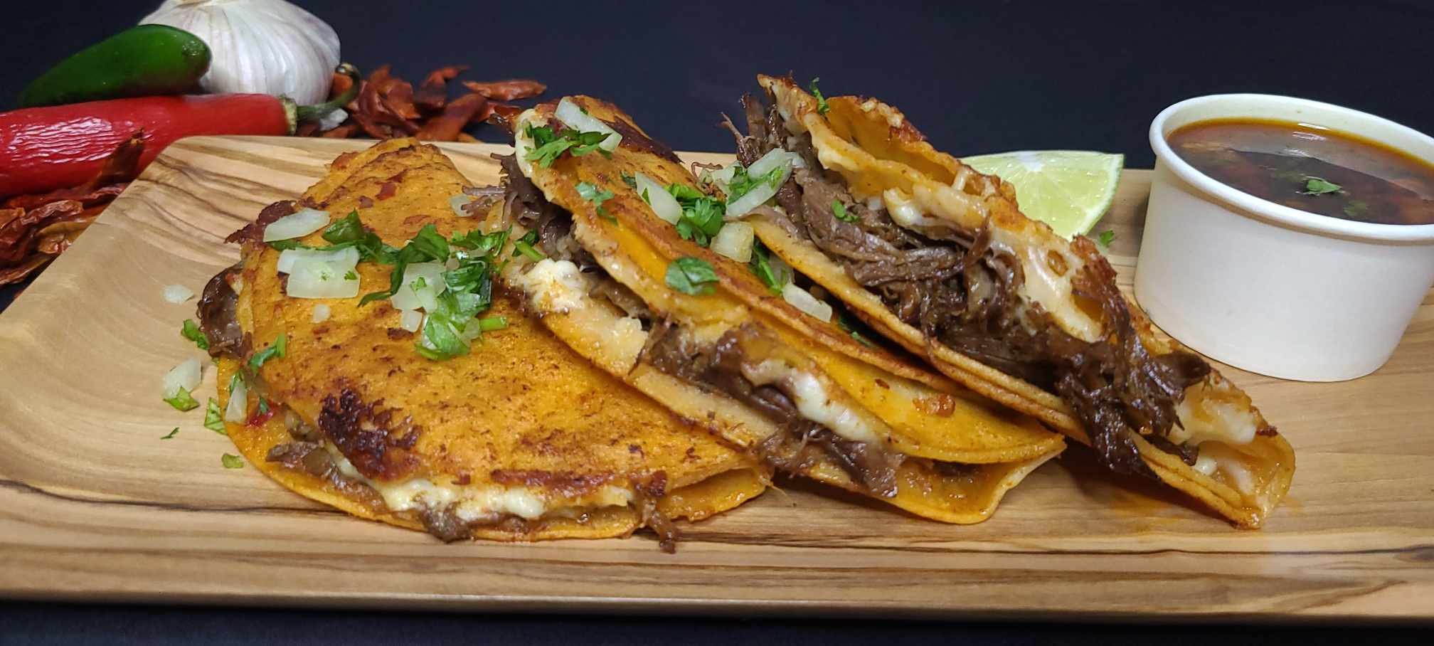 Two beef tacos with shredded beef, melted cheese, chopped onions, and cilantro, served on a wooden plate with a wedge of lime and a side of dark sauce.