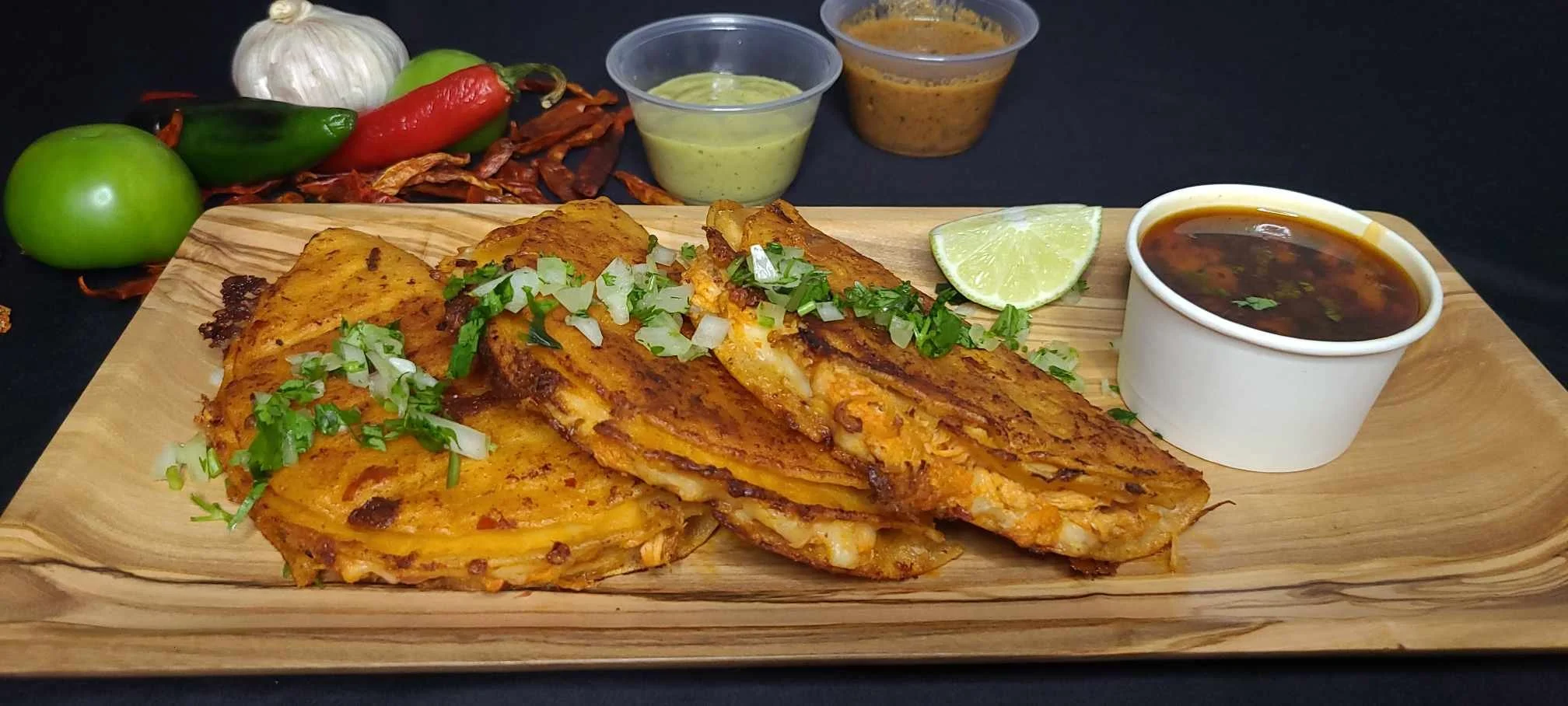 Plate with cheese quesadillas garnished with chopped onions and cilantro, lime wedge, bowl of dark broth, and small cups of green and brown sauces, with tomatoes, garlic, and chili peppers in the background.