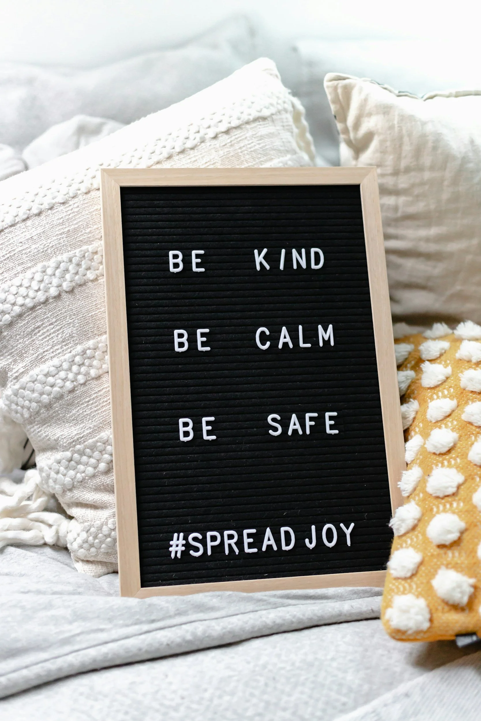 A black felt letter board with white letters displaying the message "Be kind, Be calm, Be safe, #SpreadJoy" placed on a bed with cream and beige pillows and a blanket.