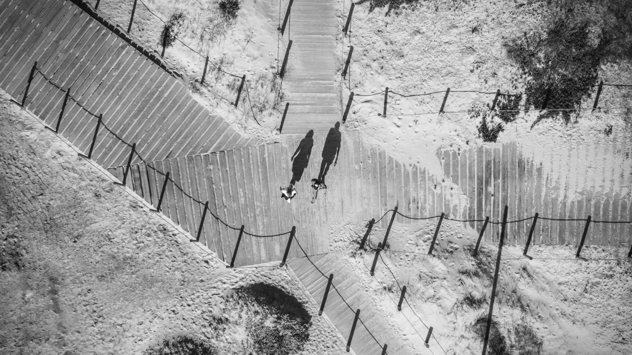 Black and white aerial photograph of two people walking on a wooden pathway through a snowy landscape, with other pathways visible in the background.