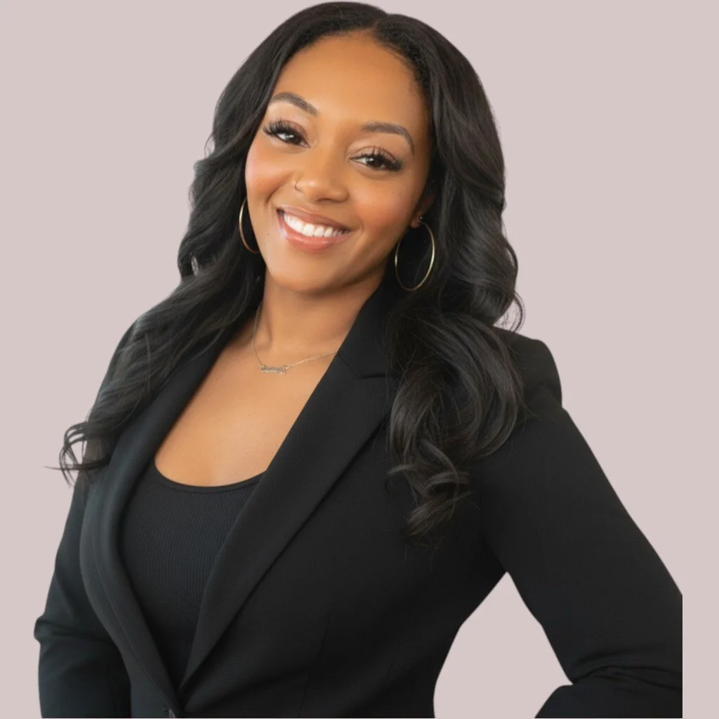 A woman with long, wavy black hair smiling, wearing a black blazer and hoop earrings, against a neutral background.