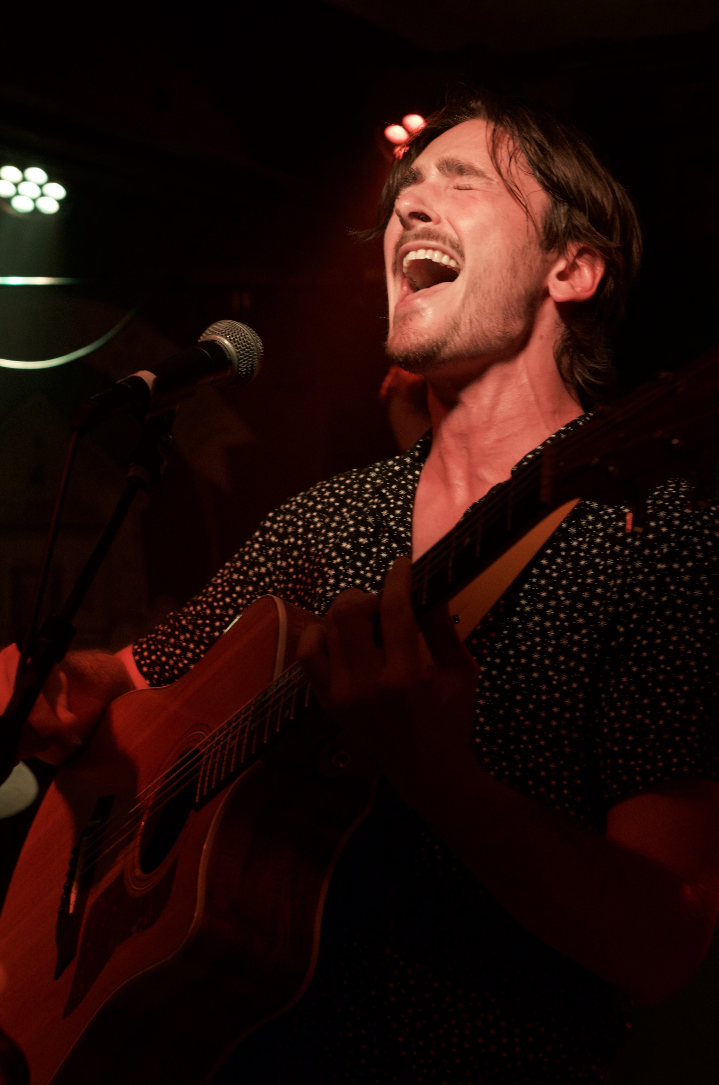 Man singing passionately while playing an acoustic guitar on stage in a dark setting.