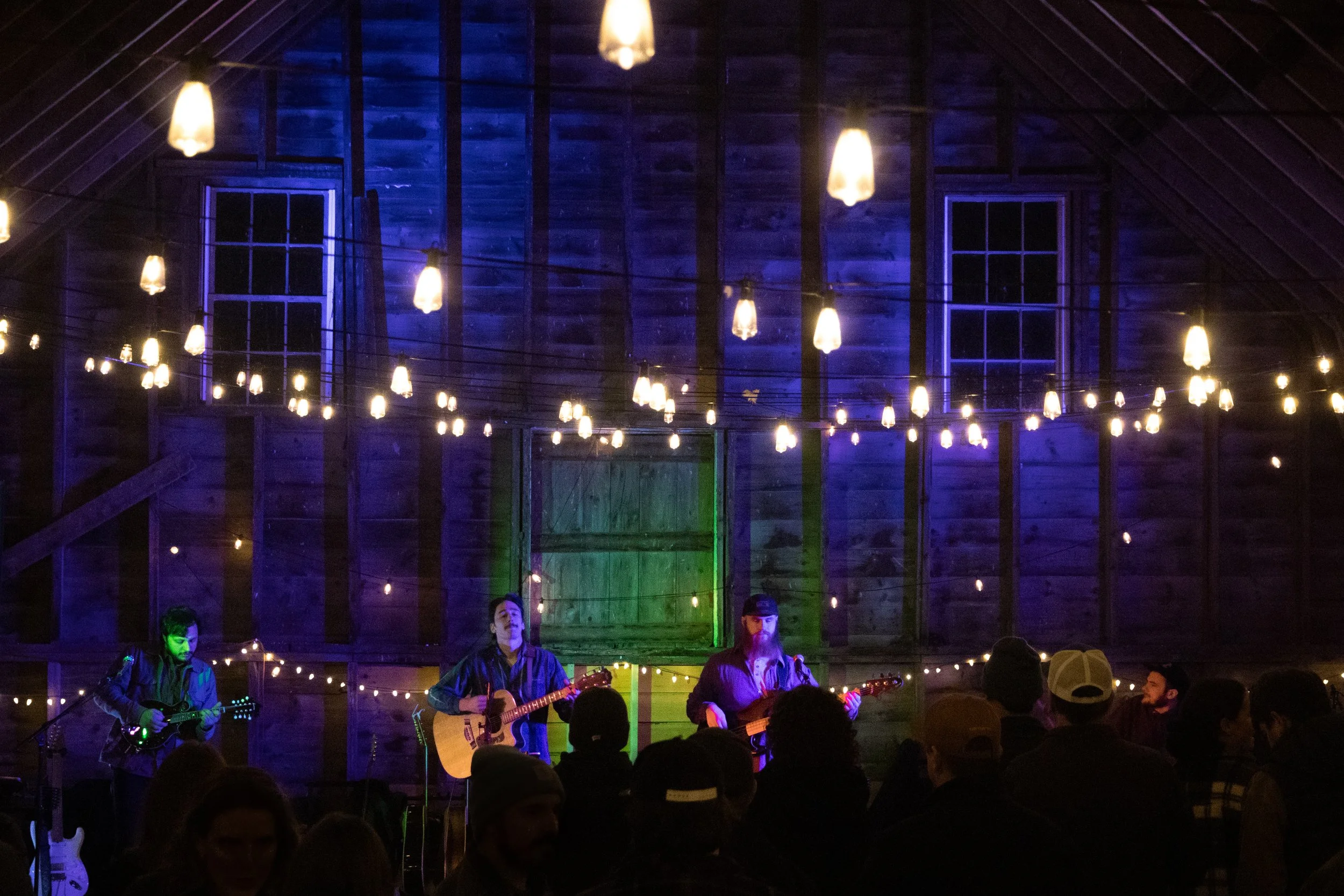Musicians perform on a stage in a rustic venue with string lights and dark wood walls, with an audience watching.