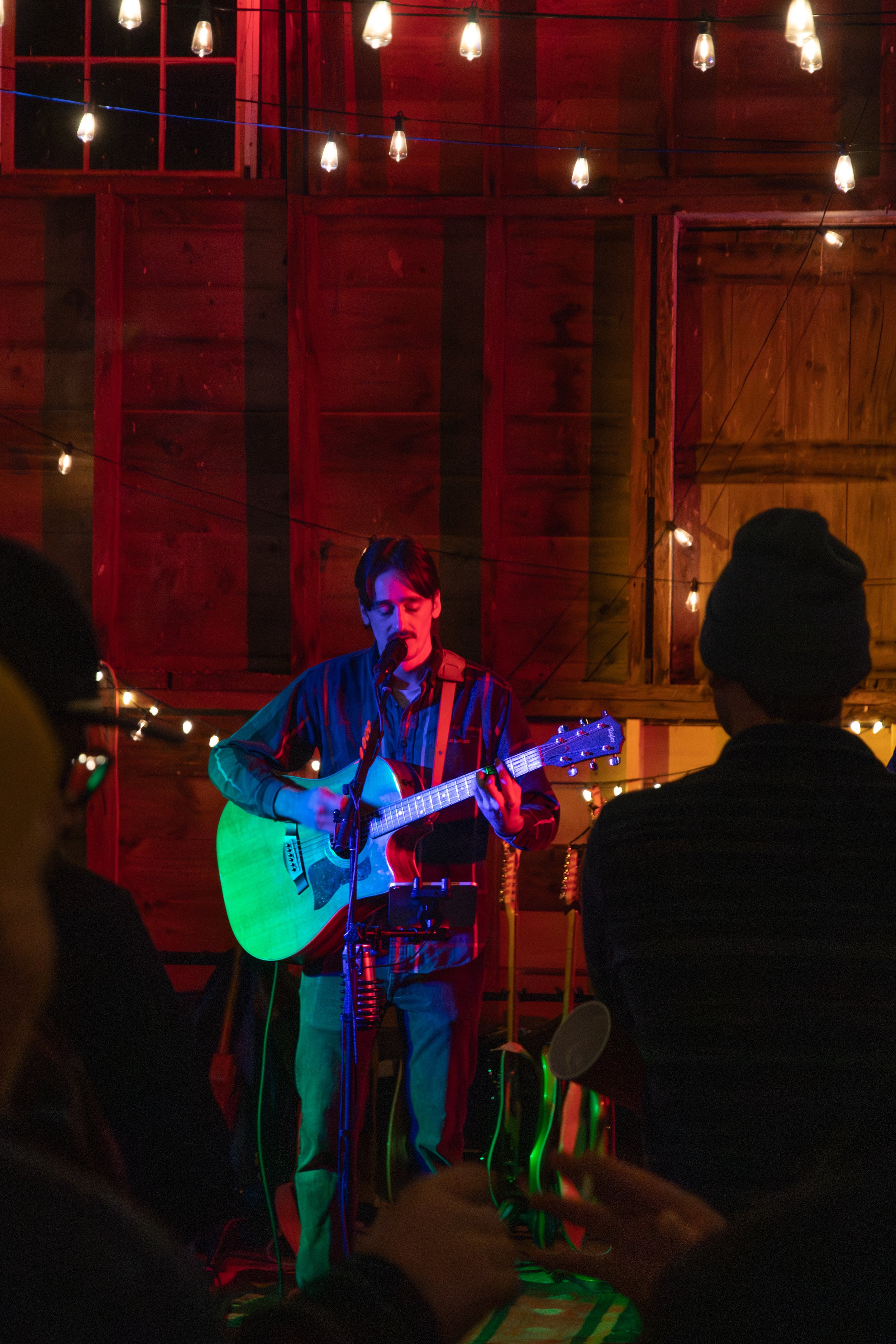 A man plays an acoustic guitar and sings into a microphone on a small stage, with string lights hanging overhead. He is wearing a plaid shirt and jeans, performing in a cozy venue with a wooden interior.