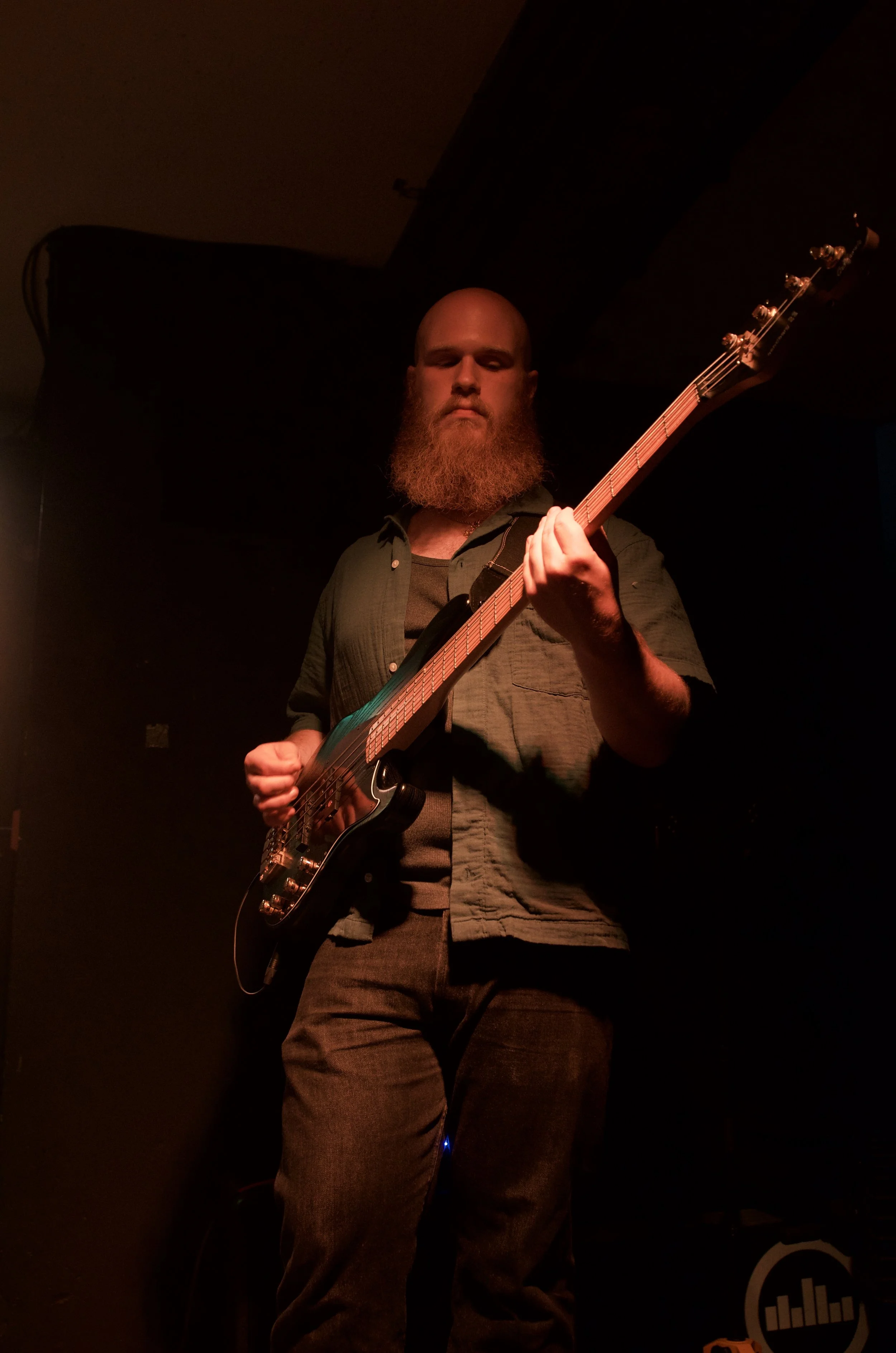 A man with a beard holding an electric bass guitar on stage in a dimly lit environment, with orange and red lighting