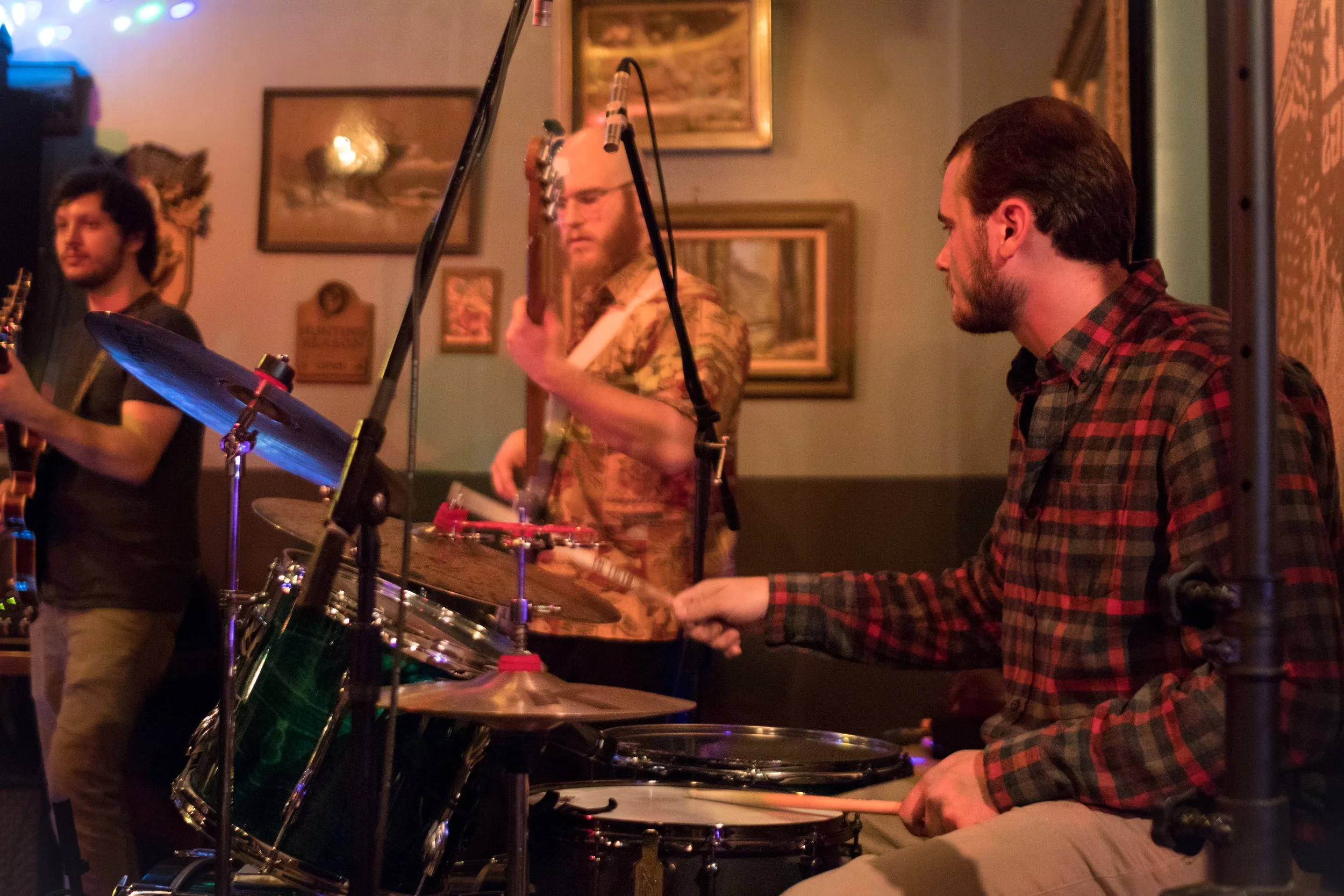 Three musicians performing indoors: a guitarist on the left, a bassist in the center, and a drummer on the right. The drummer is wearing a plaid shirt, playing a drum set, and the environment appears to be a cozy venue with framed pictures on the wal