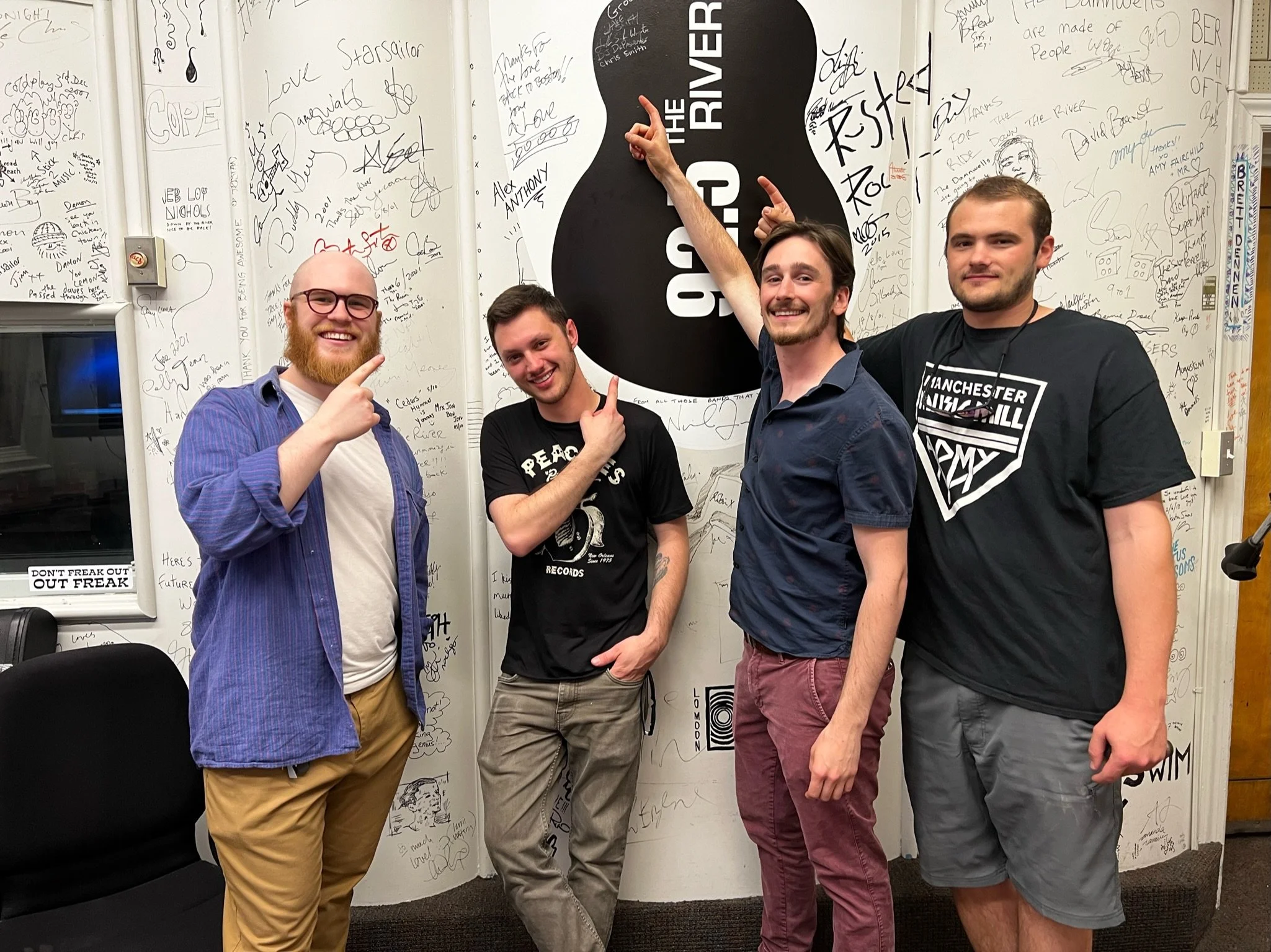Four young men standing in front of a wall covered with signatures and drawings, smiling and pointing at a large black and white sign that reads 'The River.' One man has a beard and glasses, others are smiling and gesturing, all casually dressed.