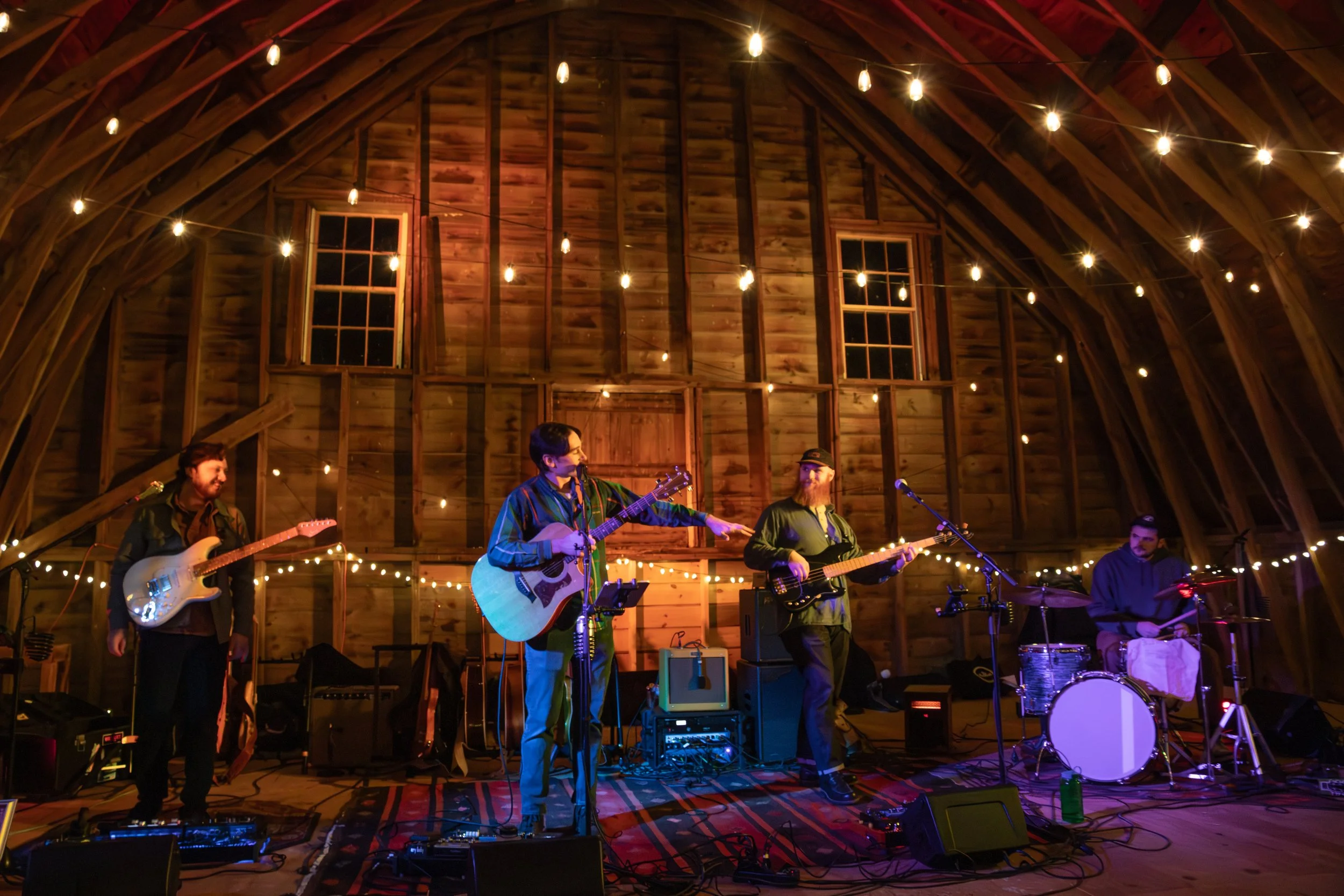 A band of five musicians performing on a stage in a rustic, wooden barn decorated with string lights.